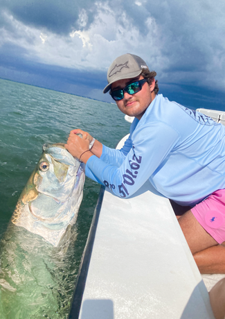 Man on a boat holding a large silver tarpon fish near the water's surface under a cloudy sky. He's wearing sunglasses, a hat, and light blue shirt.