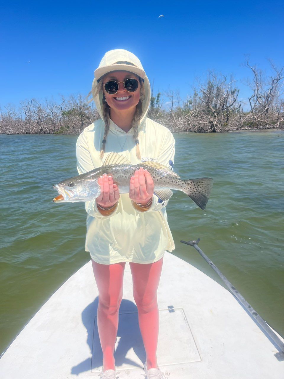 Woman on a boat smiles, holding a speckled trout. She wears a hat, sunglasses, yellow outfit, and pink leggings, set against a water backdrop.