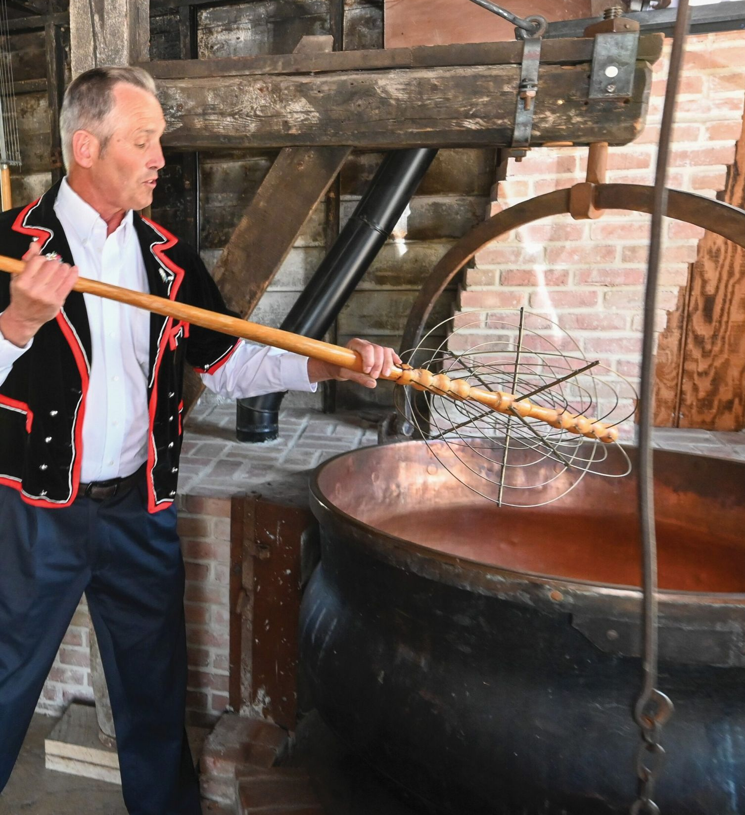 A person in traditional clothing uses a long-handled wire whisk to stir a large copper vat in a rustic, brick setting.
