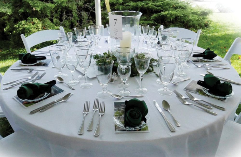 A round table set for a formal event with white tablecloths, silverware, glassware, and dark green napkins.