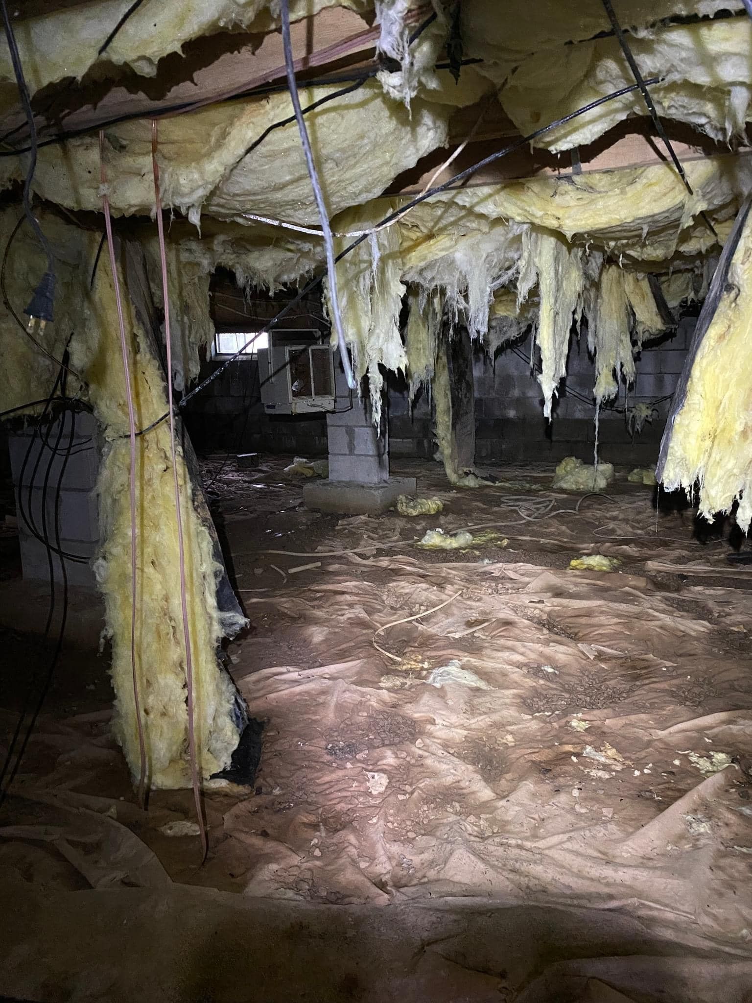 A dark crawlspace with fallen, torn yellow fiberglass insulation hanging from the ceiling joists above a dirt floor.