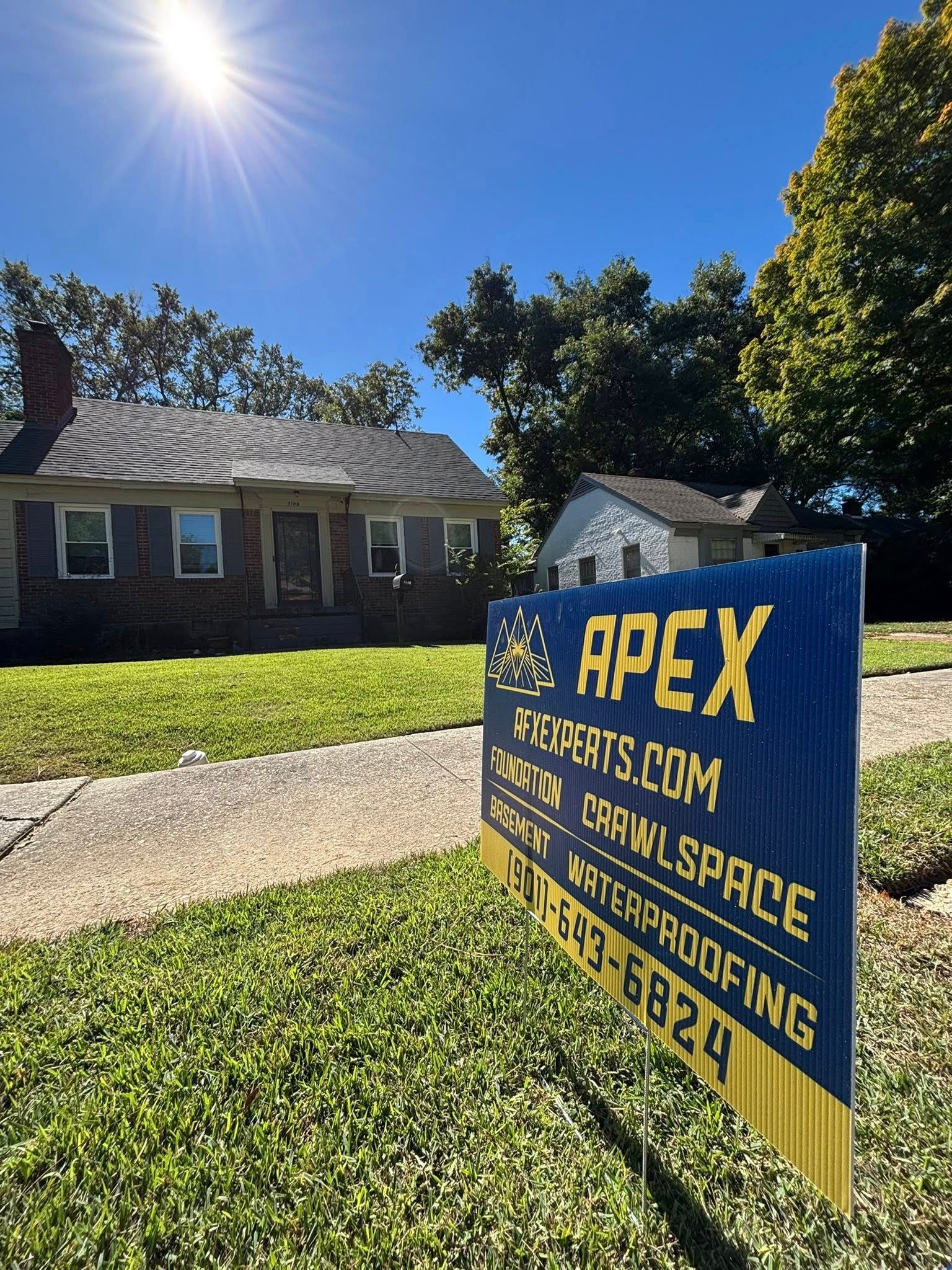 A blue and yellow Apex sign stands on a lawn in front of a brick house under a bright sunny sky.
