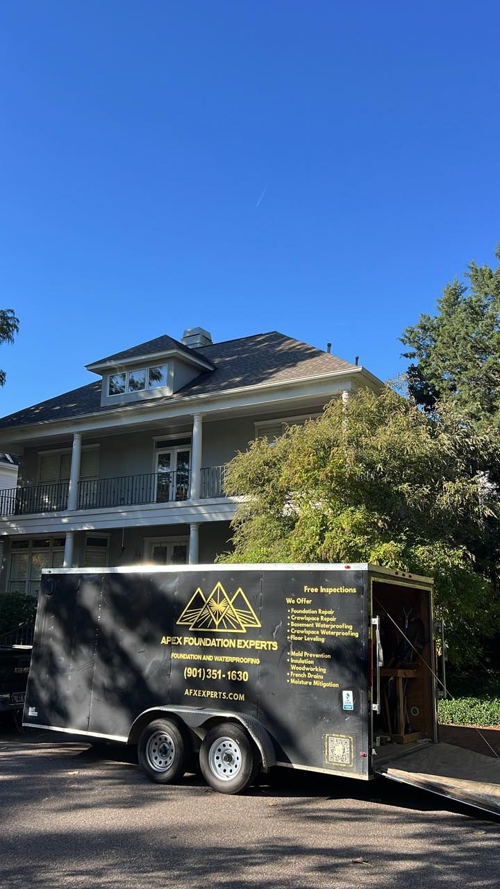 A black equipment trailer marked with gold logo and text parked in front of a two-story gray house under a clear blue sky.