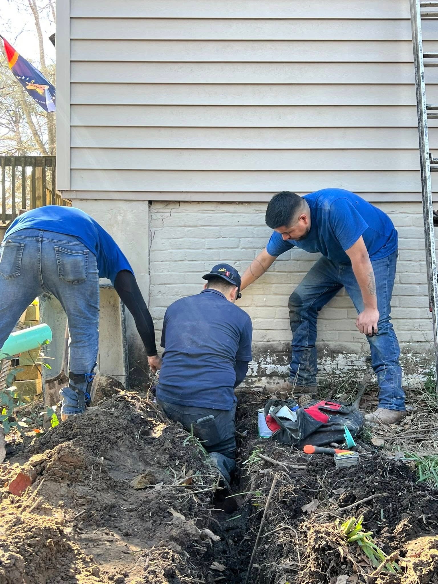 Three people in blue work uniforms work in a trench against a house foundation.