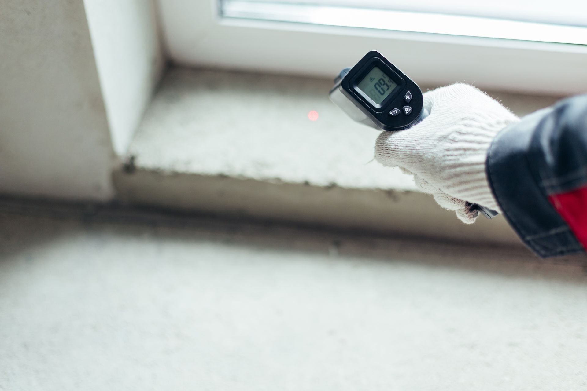 A person wearing a white work glove uses an infrared thermometer to measure the temperature of a concrete windowsill.