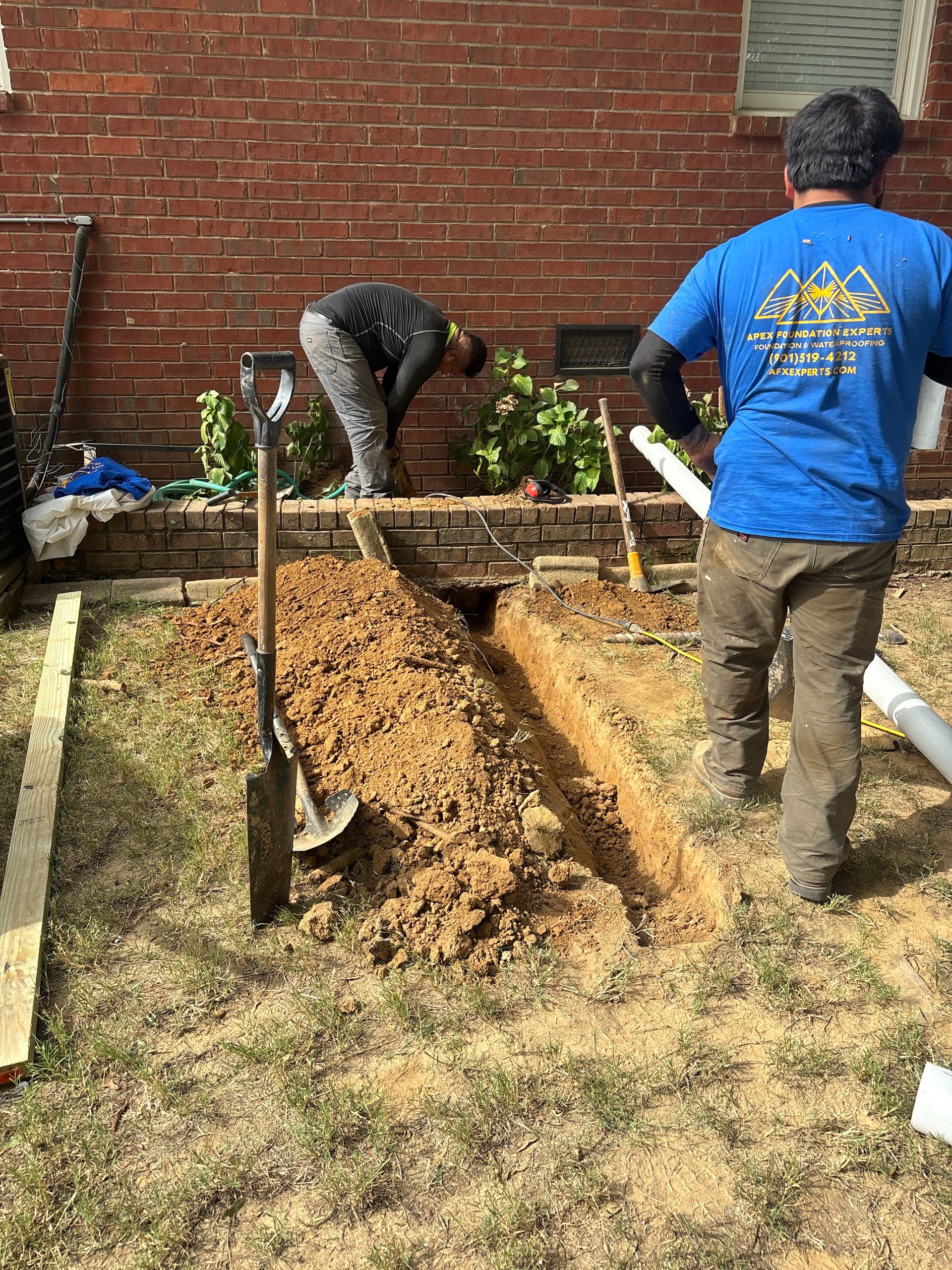 Two workers dig a trench in a yard alongside a brick house wall.