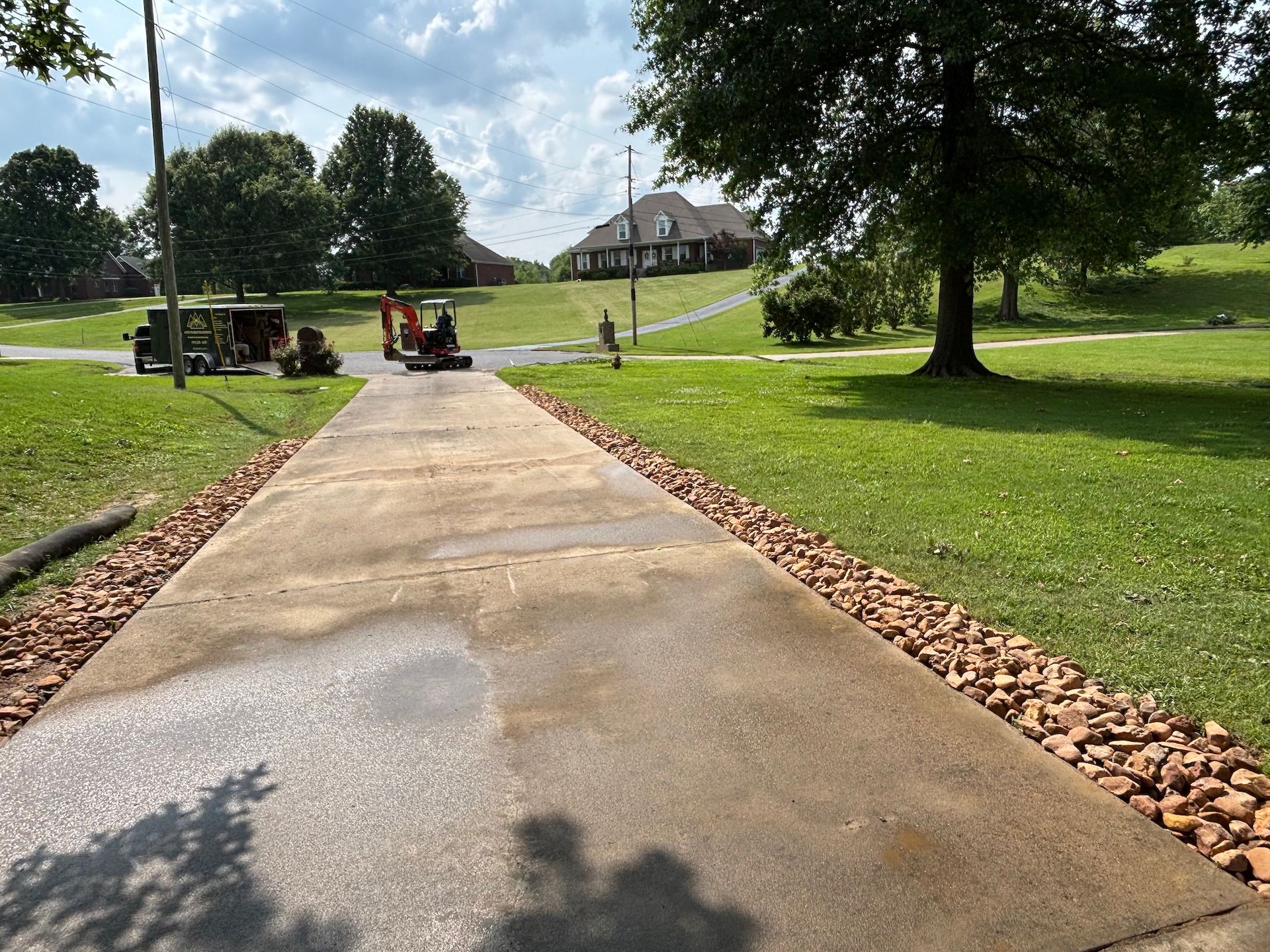A concrete driveway leads toward a house, bordered by brown landscape rocks on a sunny, green lawn.