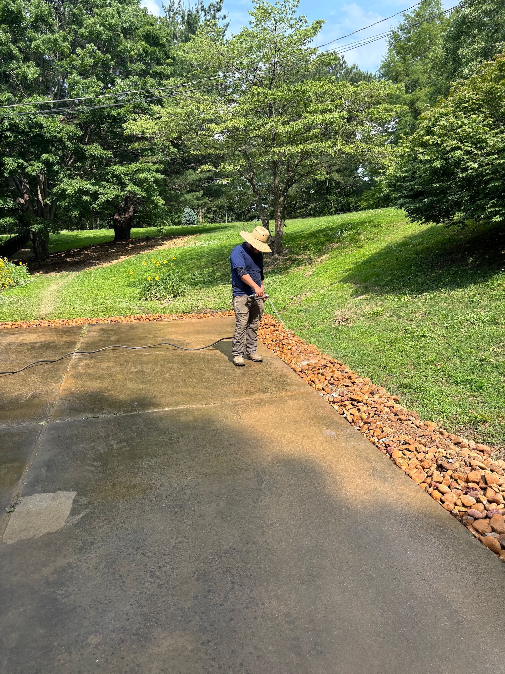 A person wearing a wide-brimmed hat stands on a paved driveway bordered by a row of decorative rocks and a grassy hill.
