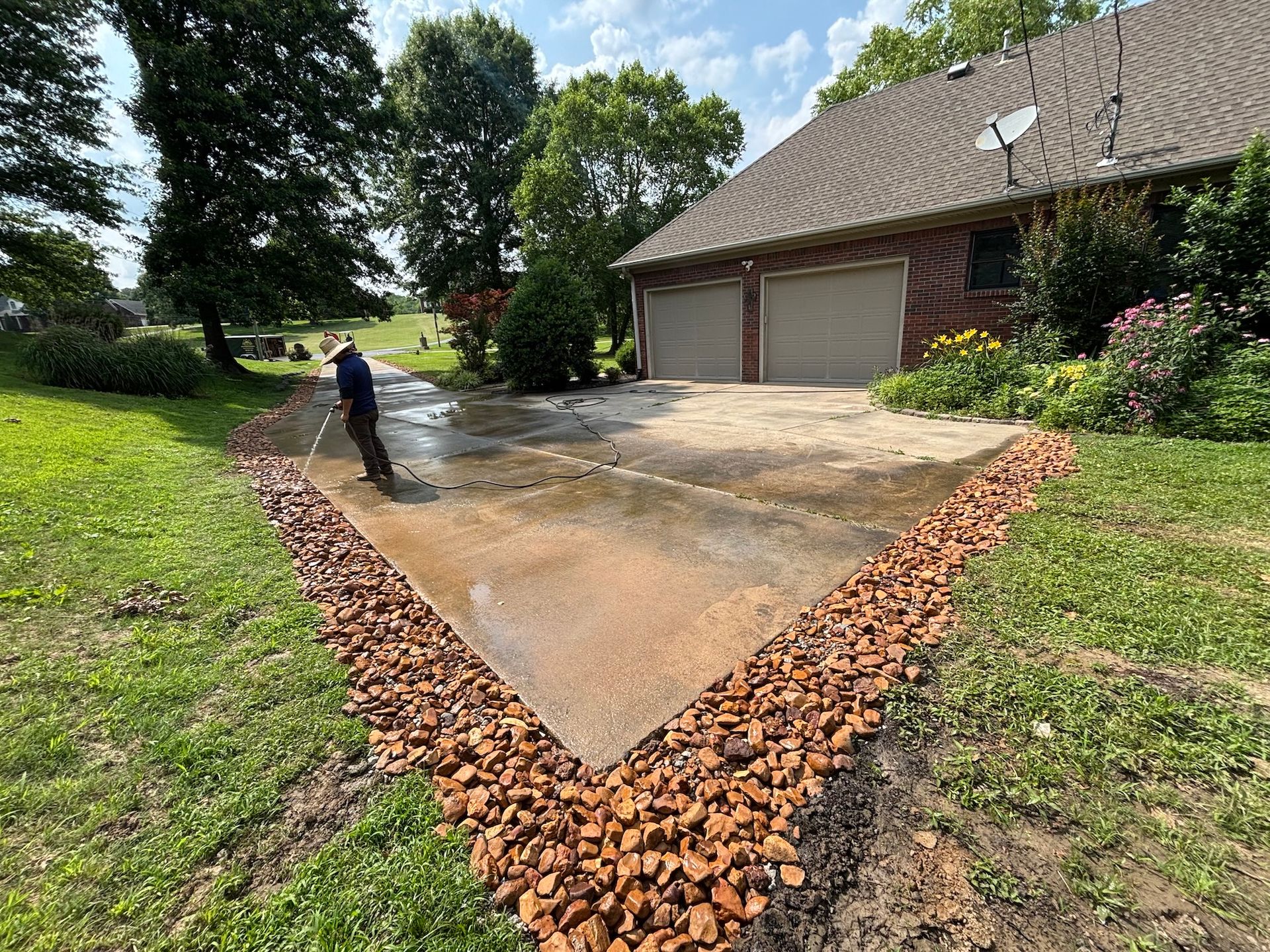 A person power-washes a concrete driveway bordering a house with rock landscaping on a sunny day.
