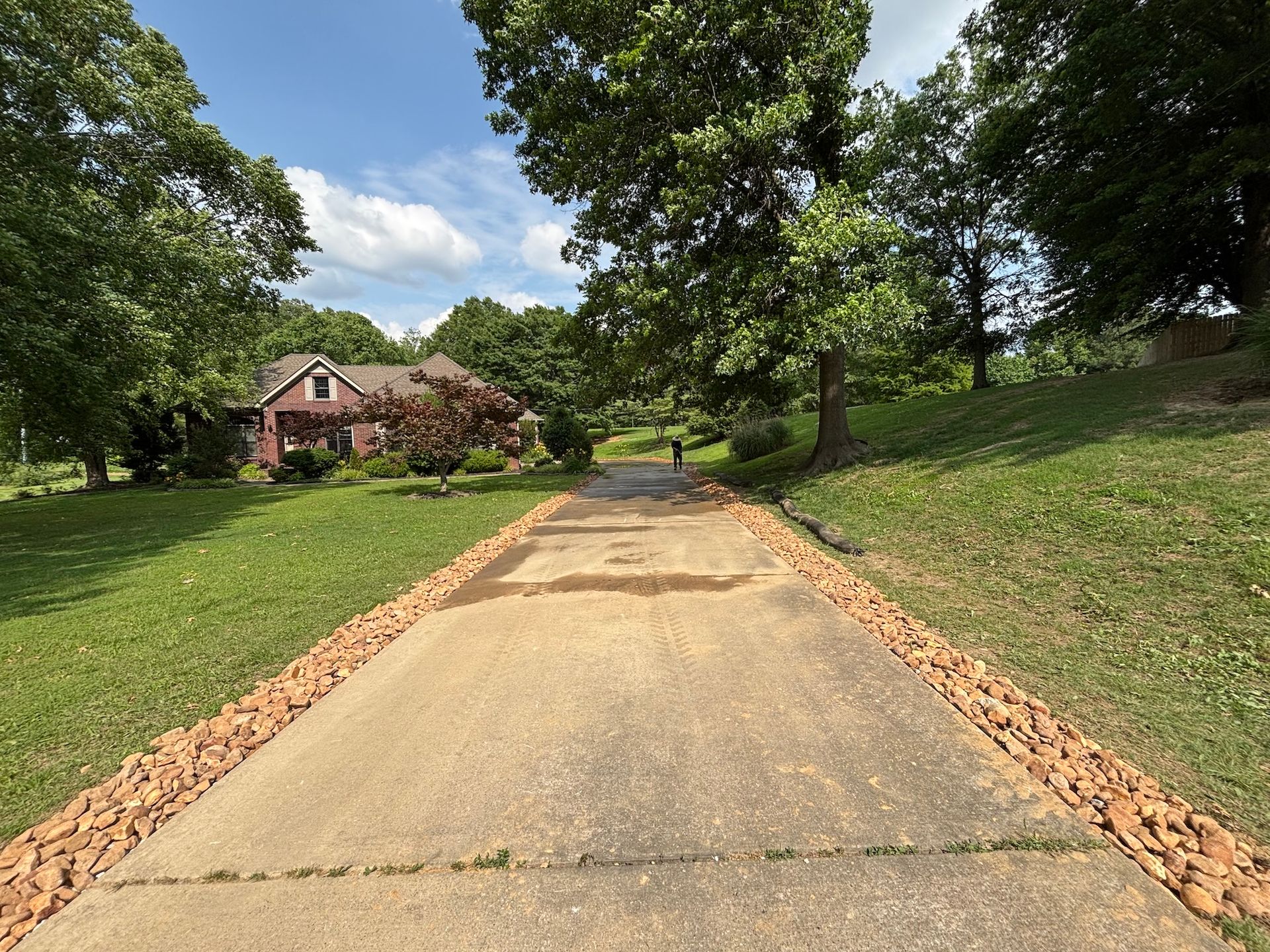A concrete driveway lined with decorative stones leads toward a house surrounded by trees and a green lawn under a blue sky.