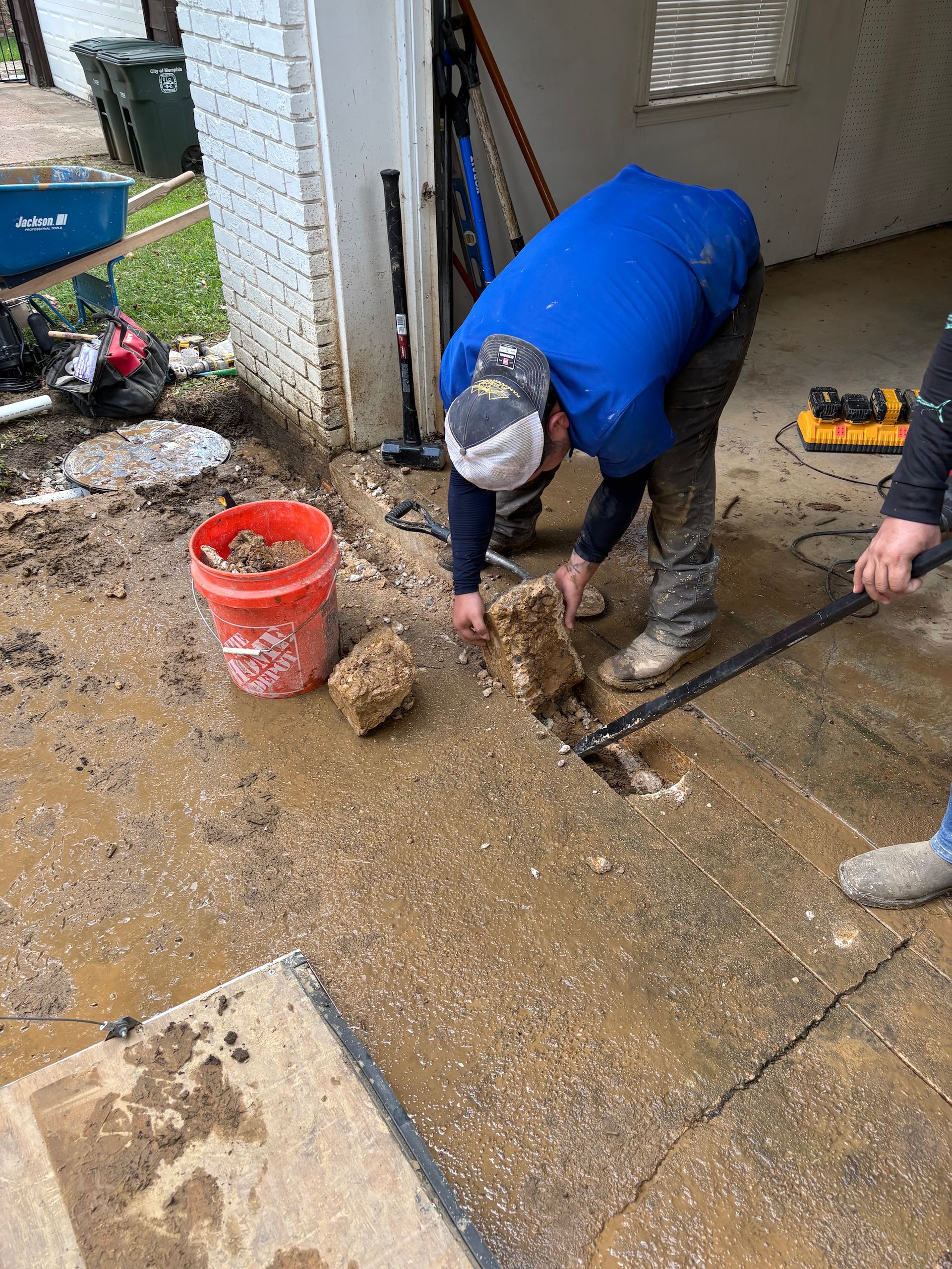 A person in a blue shirt works on an outdoor concrete patch, using a pry bar to remove debris near an orange bucket.