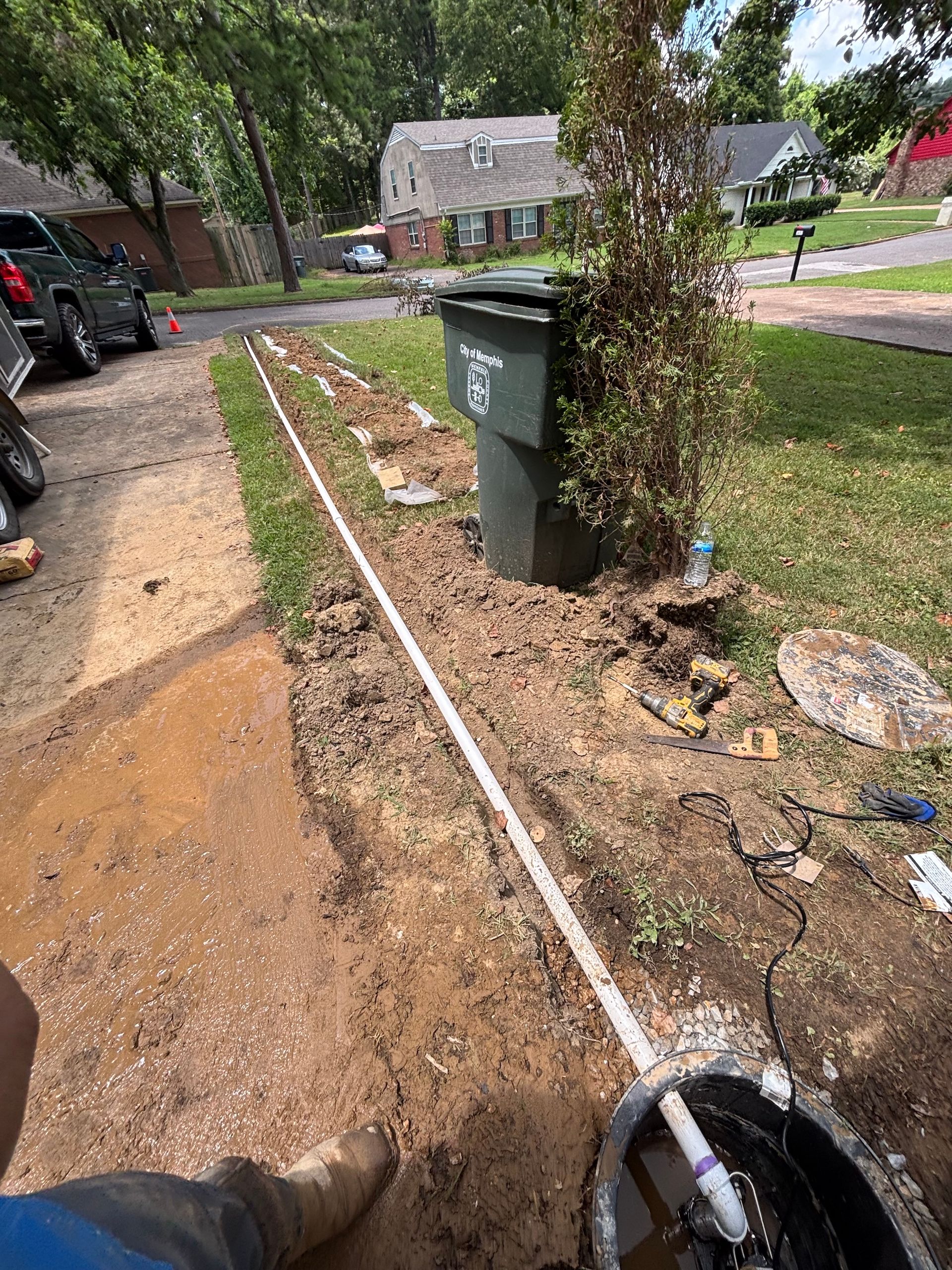 A white PVC pipe runs along a dirt trench in a residential front yard near a driveway and a green trash bin.