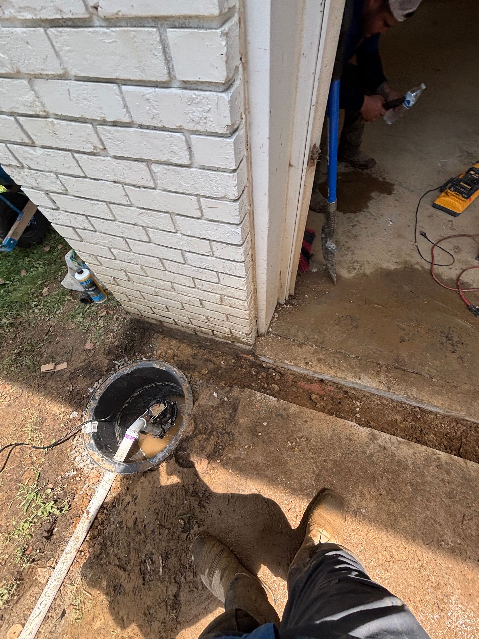 A construction worker uses a tool near a white brick wall and an open doorway where a circular basin sits in the dirt.