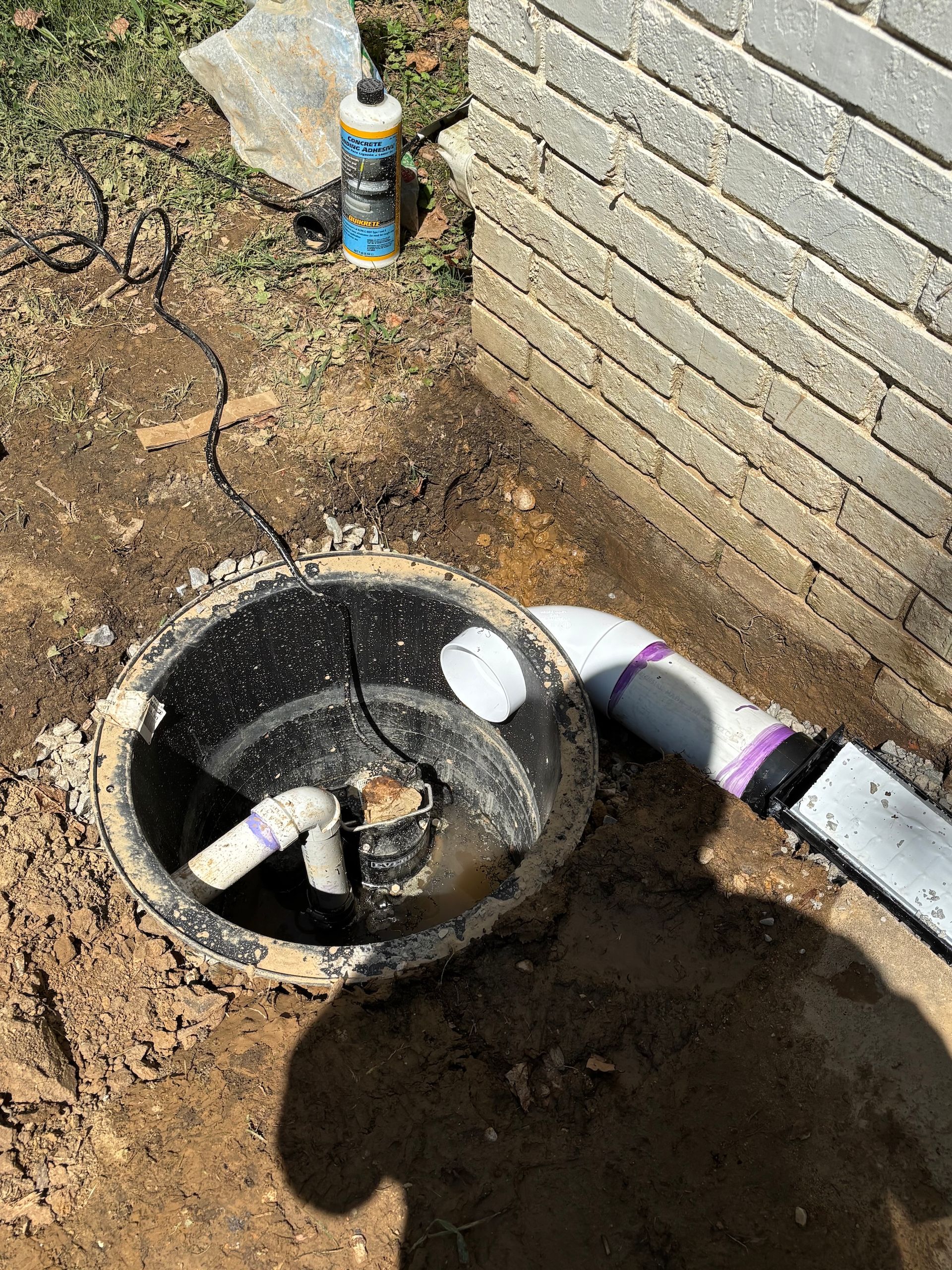 A sump pump basin installed in the ground next to a brick wall, with a PVC discharge pipe leading away from the basin.