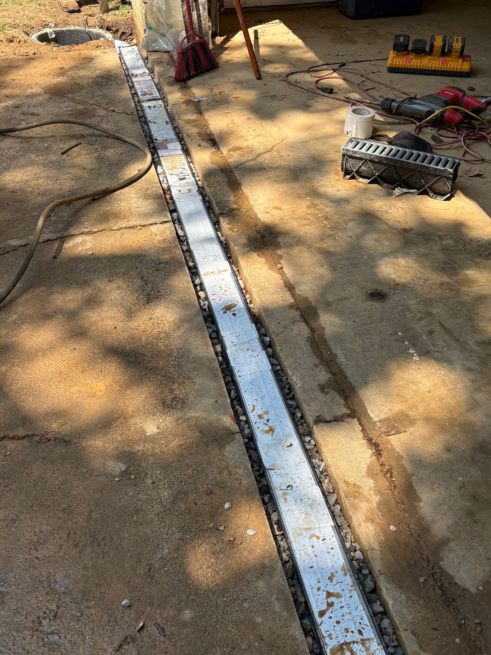 A metal trench drain is being installed in a concrete driveway, with construction tools nearby on the ground.