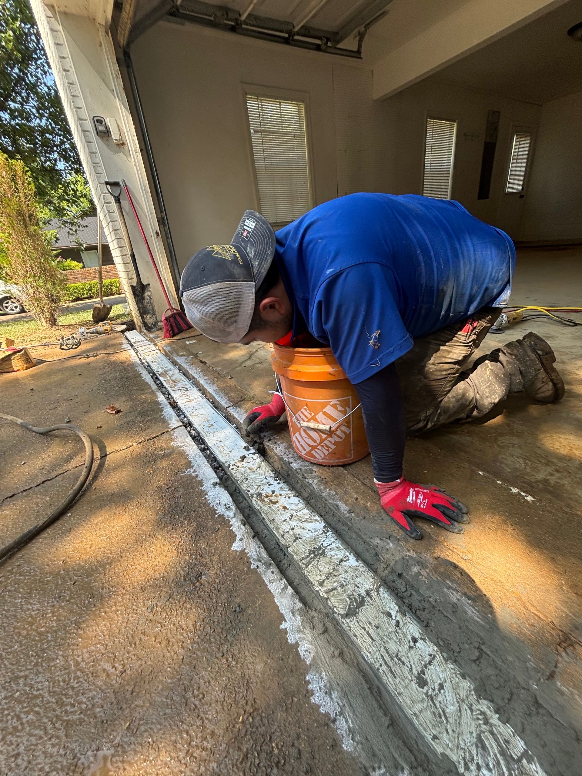 A person kneeling in a garage, using a tool to apply sealant into a floor crack, wearing a blue shirt and red gloves.