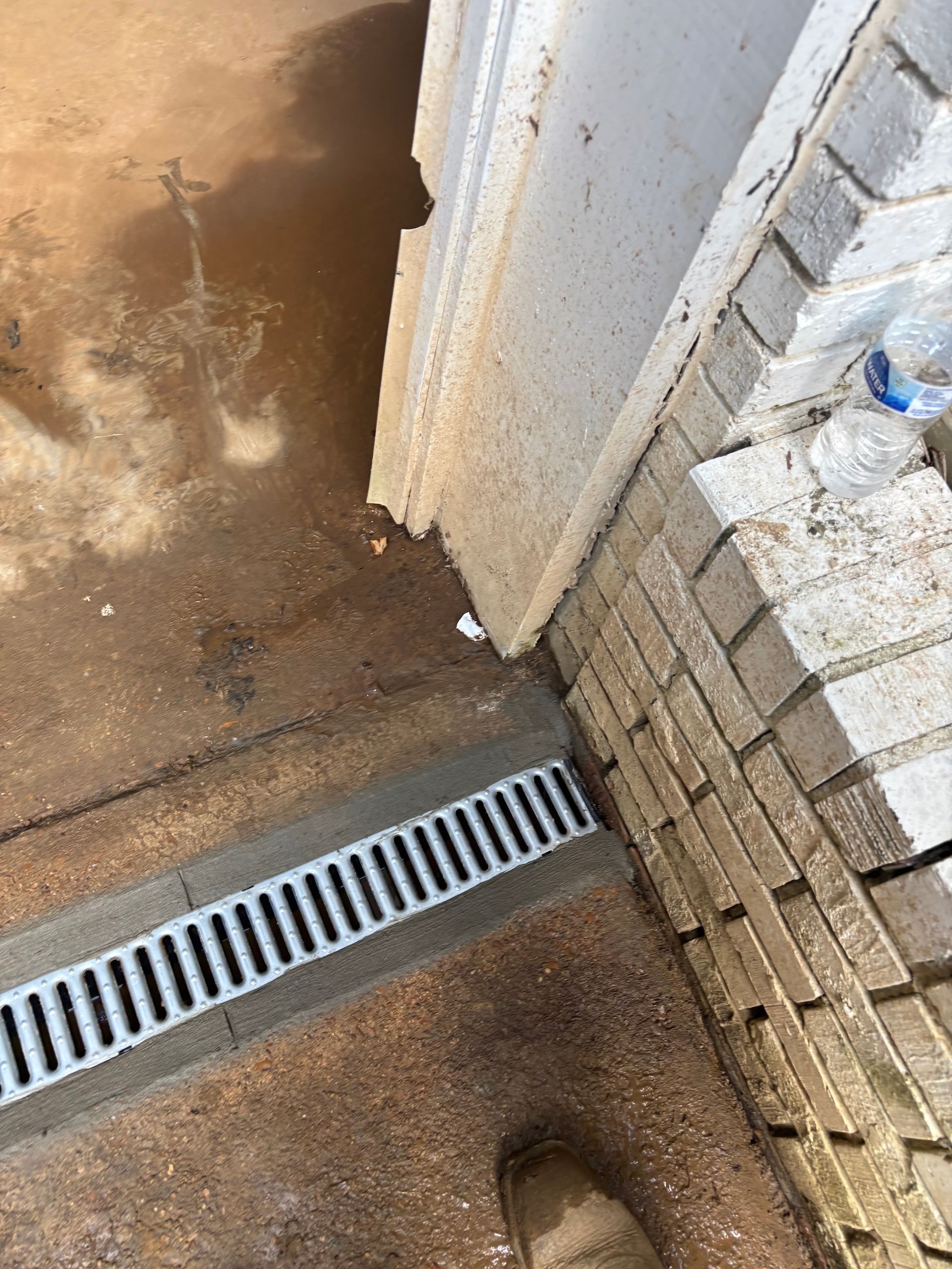 A metal trench drain installed in a dirt ground near a brick wall and a doorway, with mud visible on the surface.