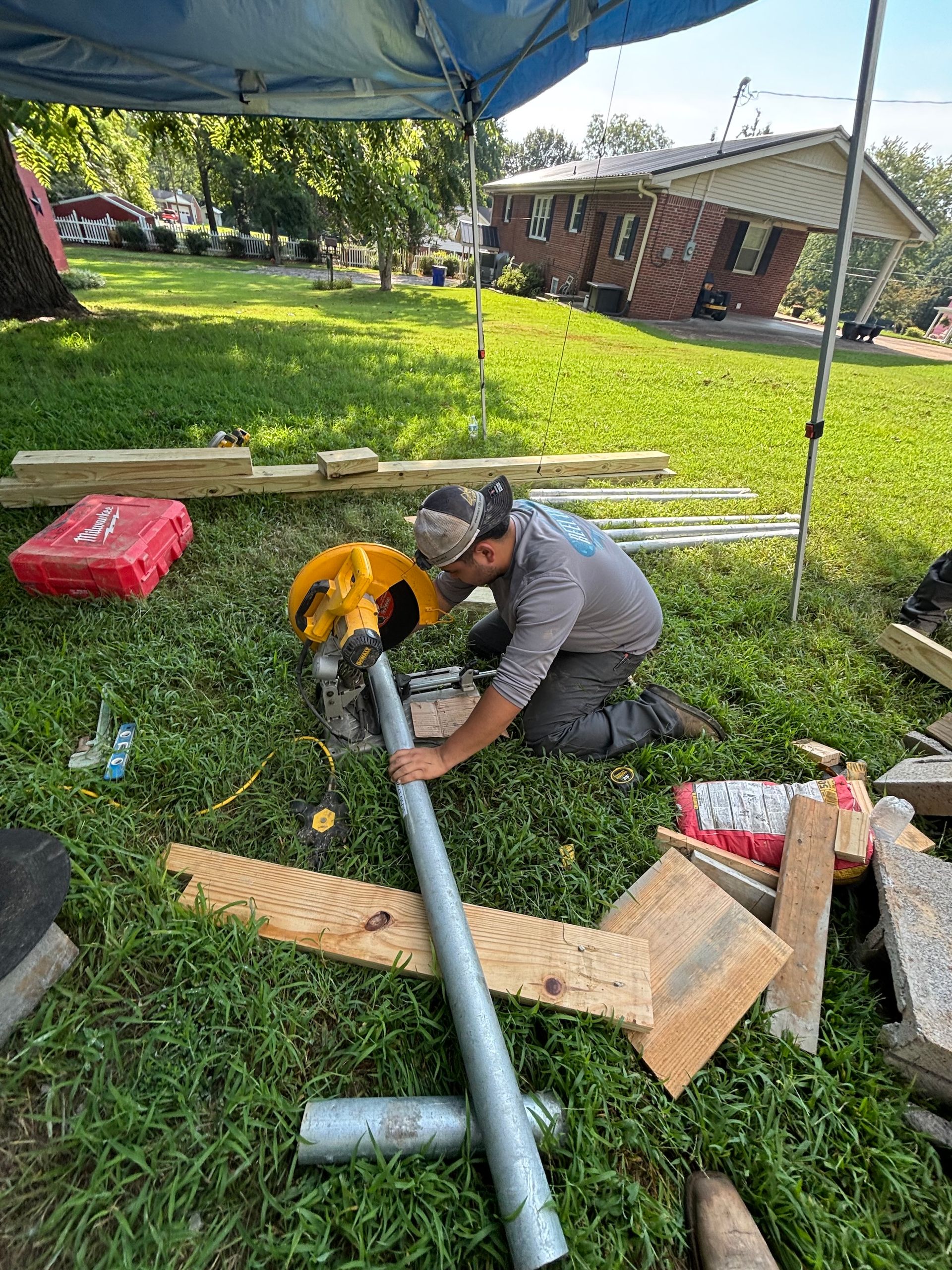 A person kneels on grass under a canopy, cutting a metal pole with a yellow circular saw.