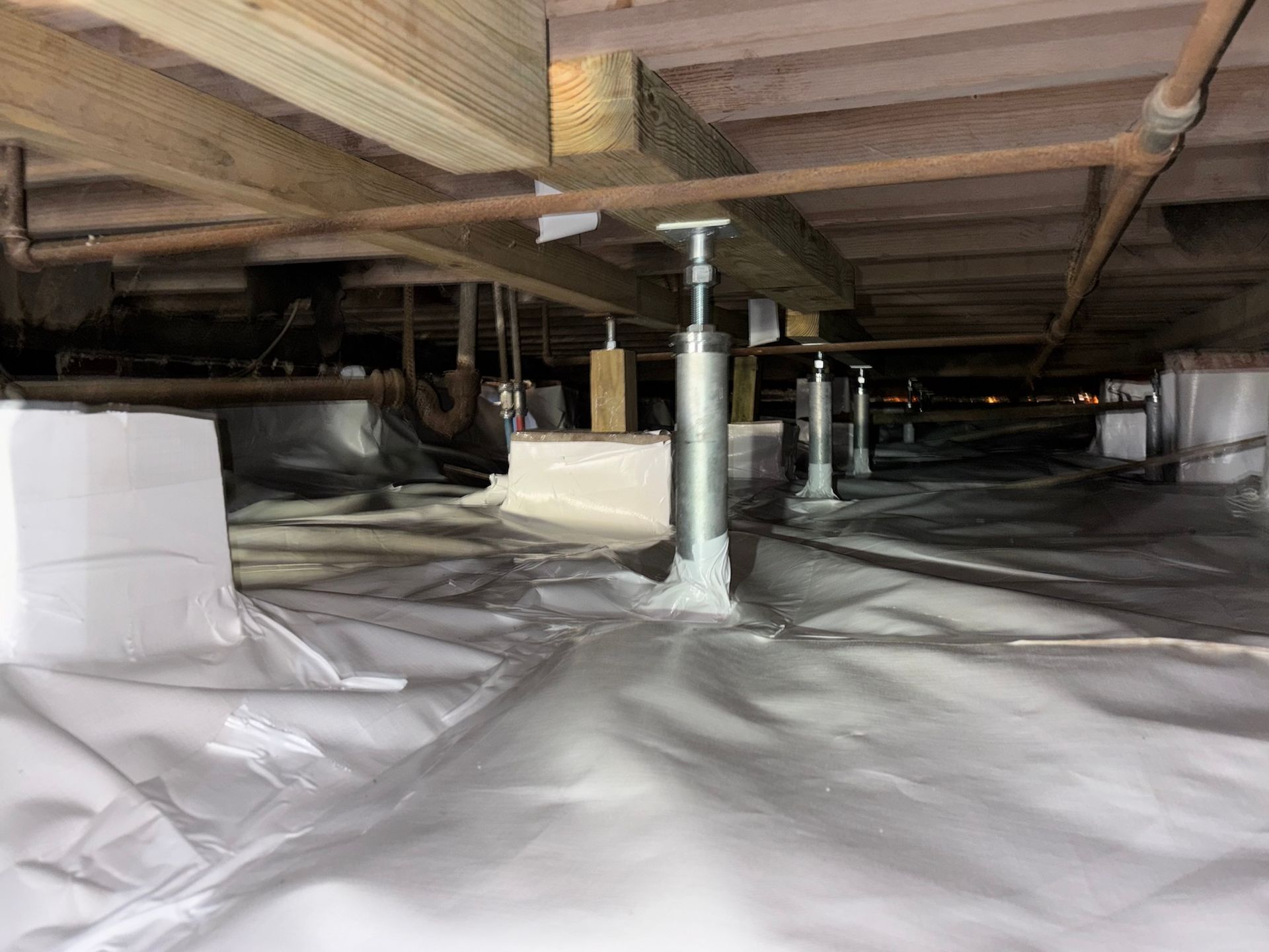 Crawl space with white vapor barrier, metal support jacks, and wooden beams under a house.