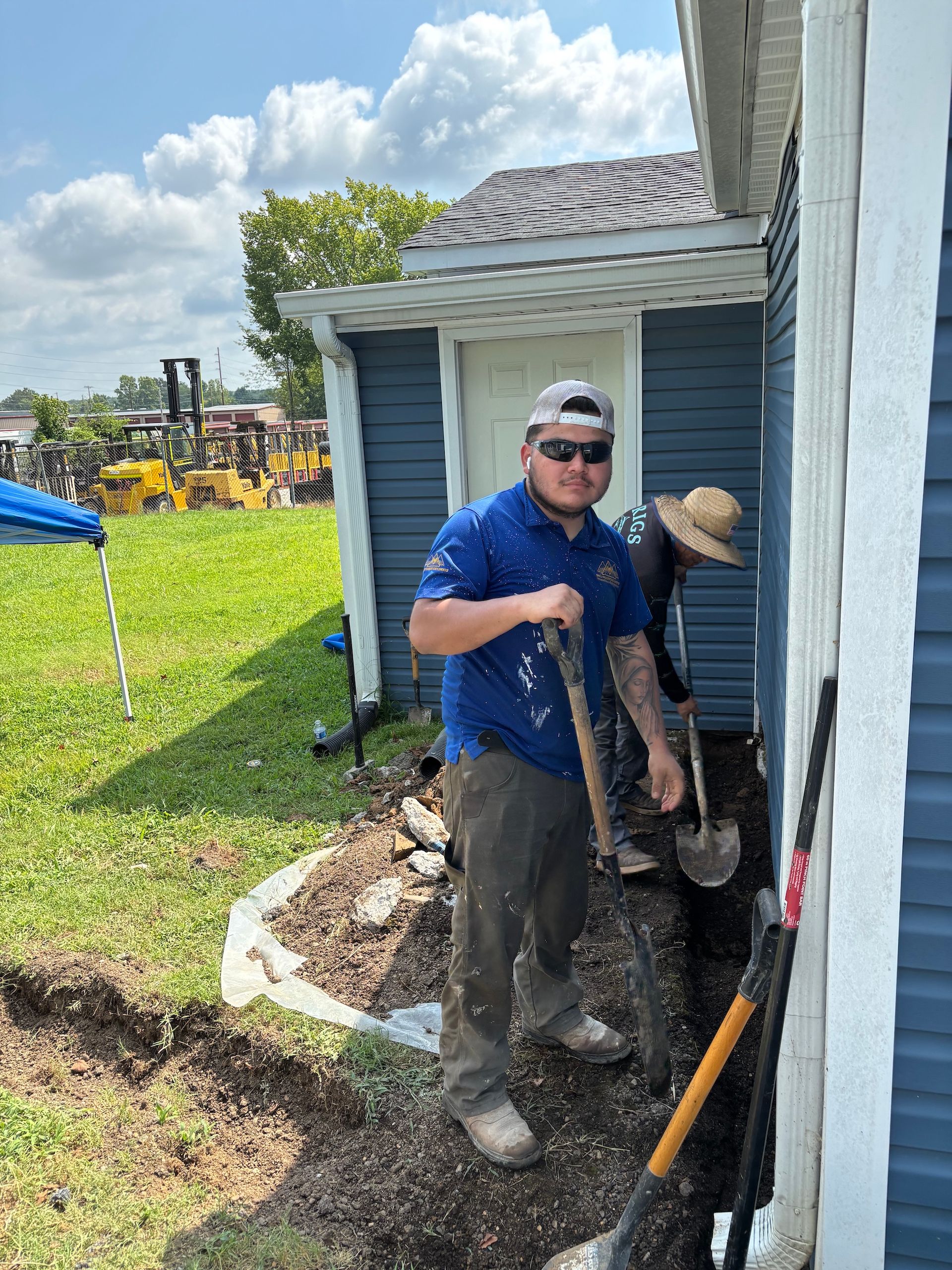 A person in a blue shirt and sunglasses uses a shovel to dig a trench along the exterior wall of a blue house.