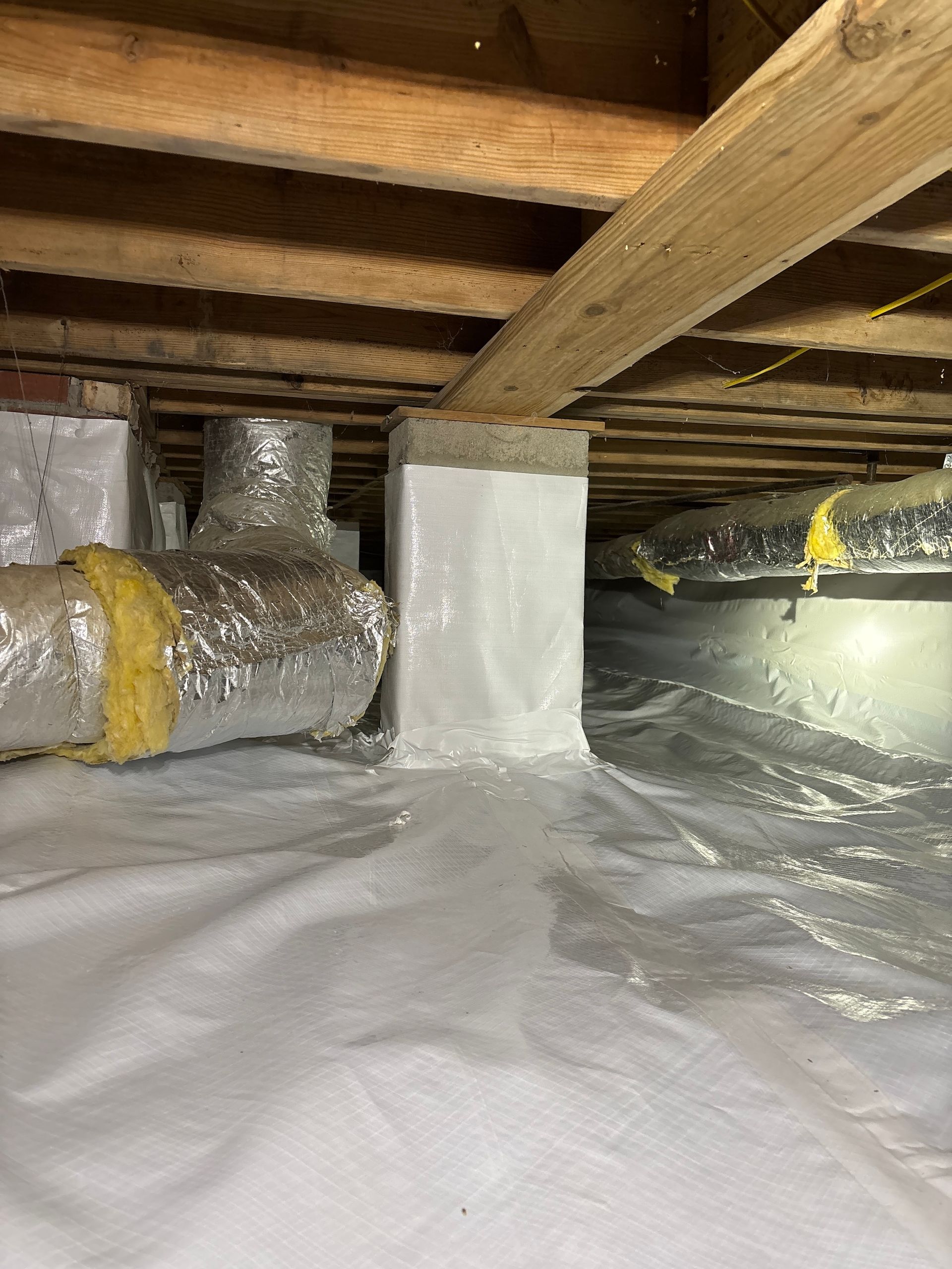 Crawl space with insulated HVAC ducts and support pillars wrapped in white vapor barrier material.