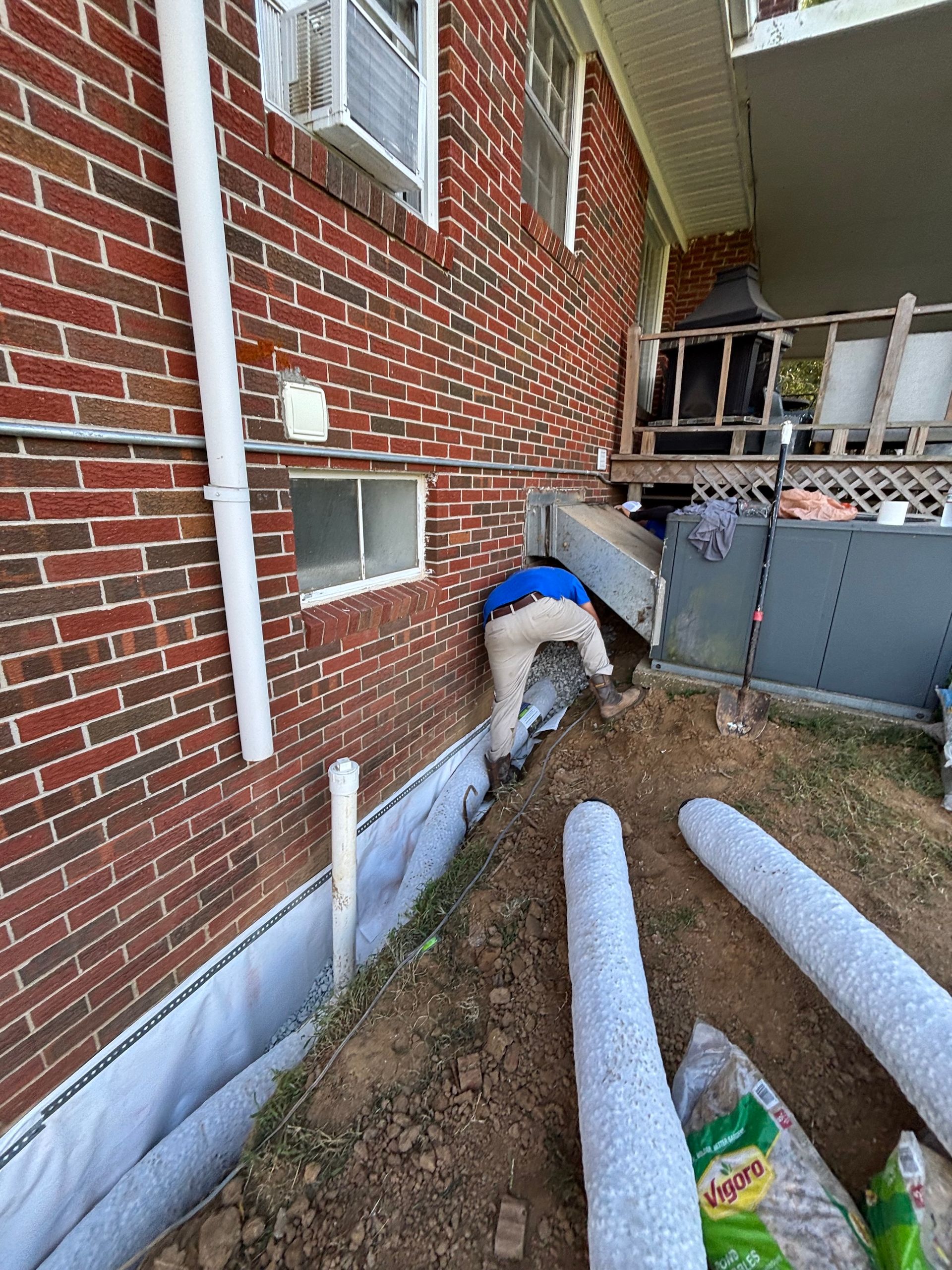 A person wearing a blue shirt works on a drainage system installation along the side of a brick house foundation.