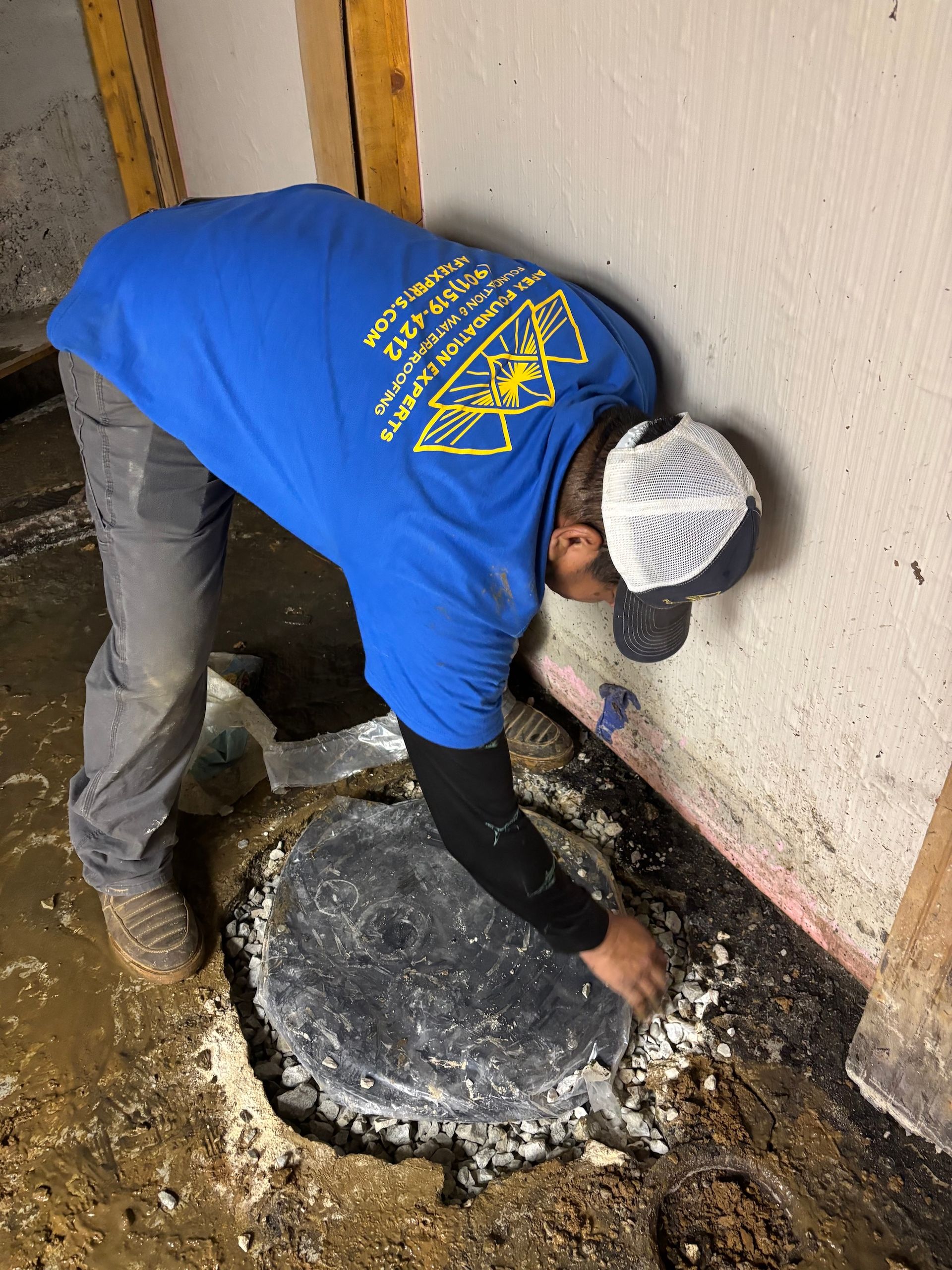 A person in a blue shirt works on a circular concrete patch in a dirt floor near a wall.