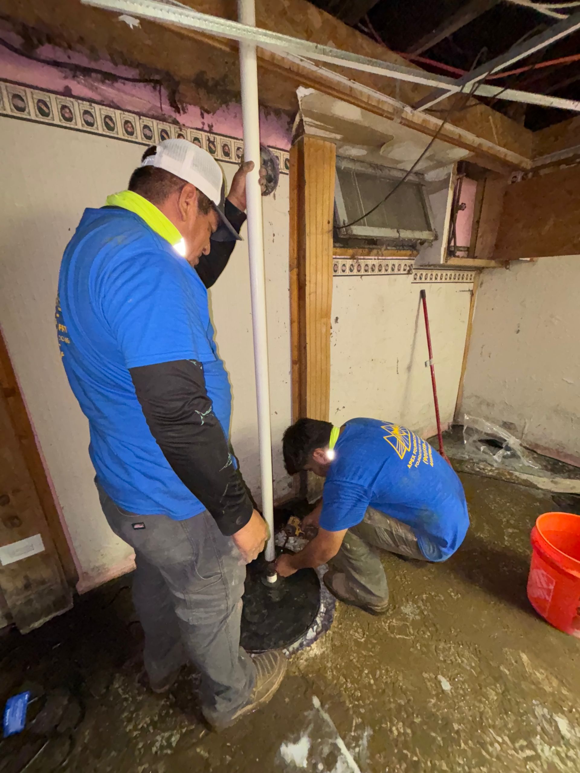 Two workers in blue shirts install a vertical pipe into a concrete floor in an unfinished basement.