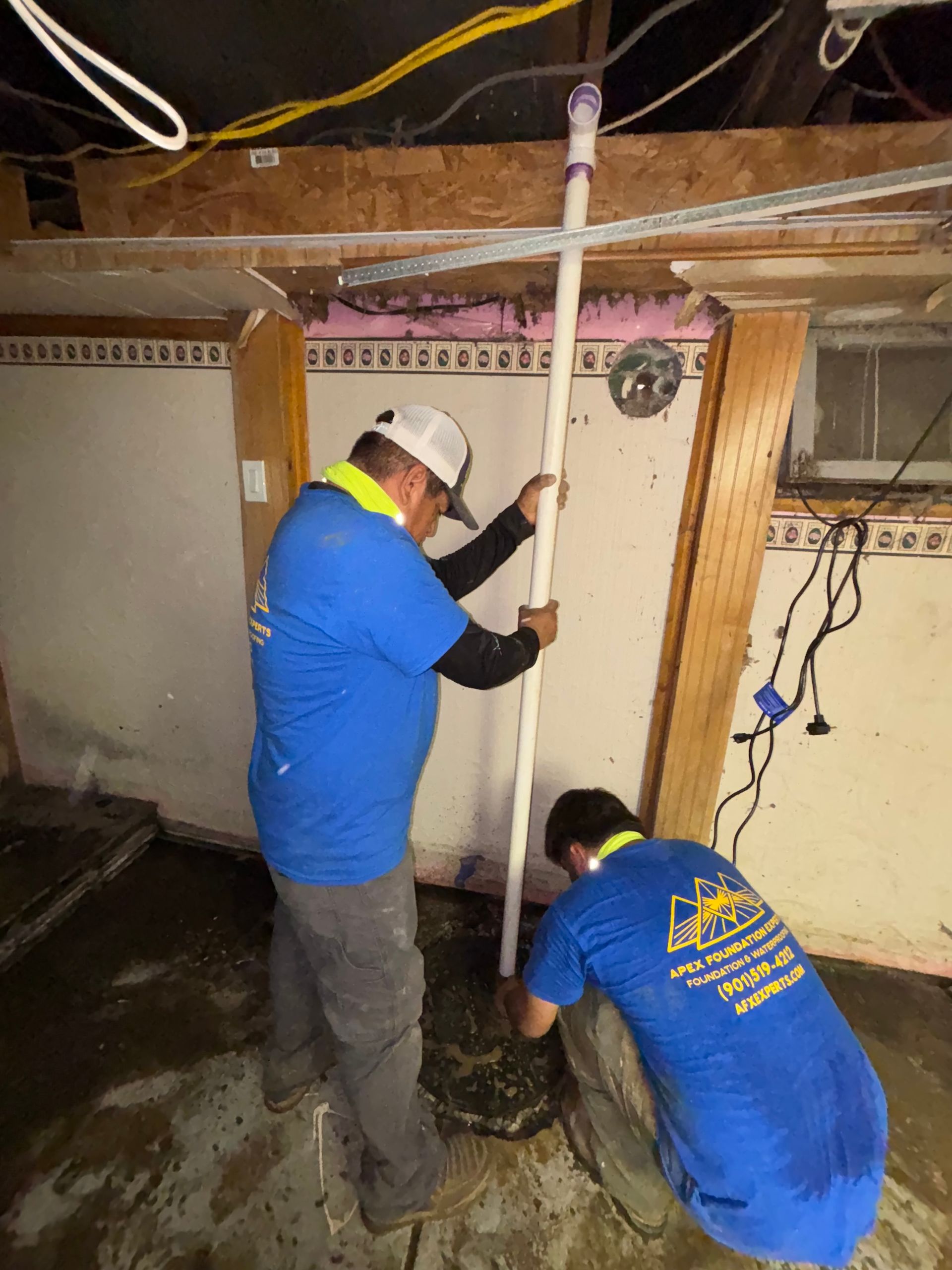 Two workers in blue uniforms install a vertical PVC pipe into a concrete basement floor.