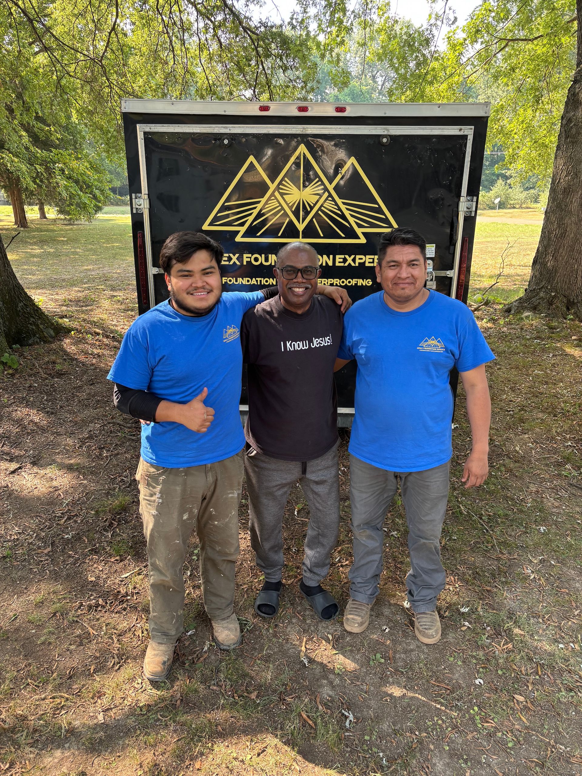 Three smiling men stand in front of a trailer with a gold triangular logo, positioned outdoors among trees and grass.