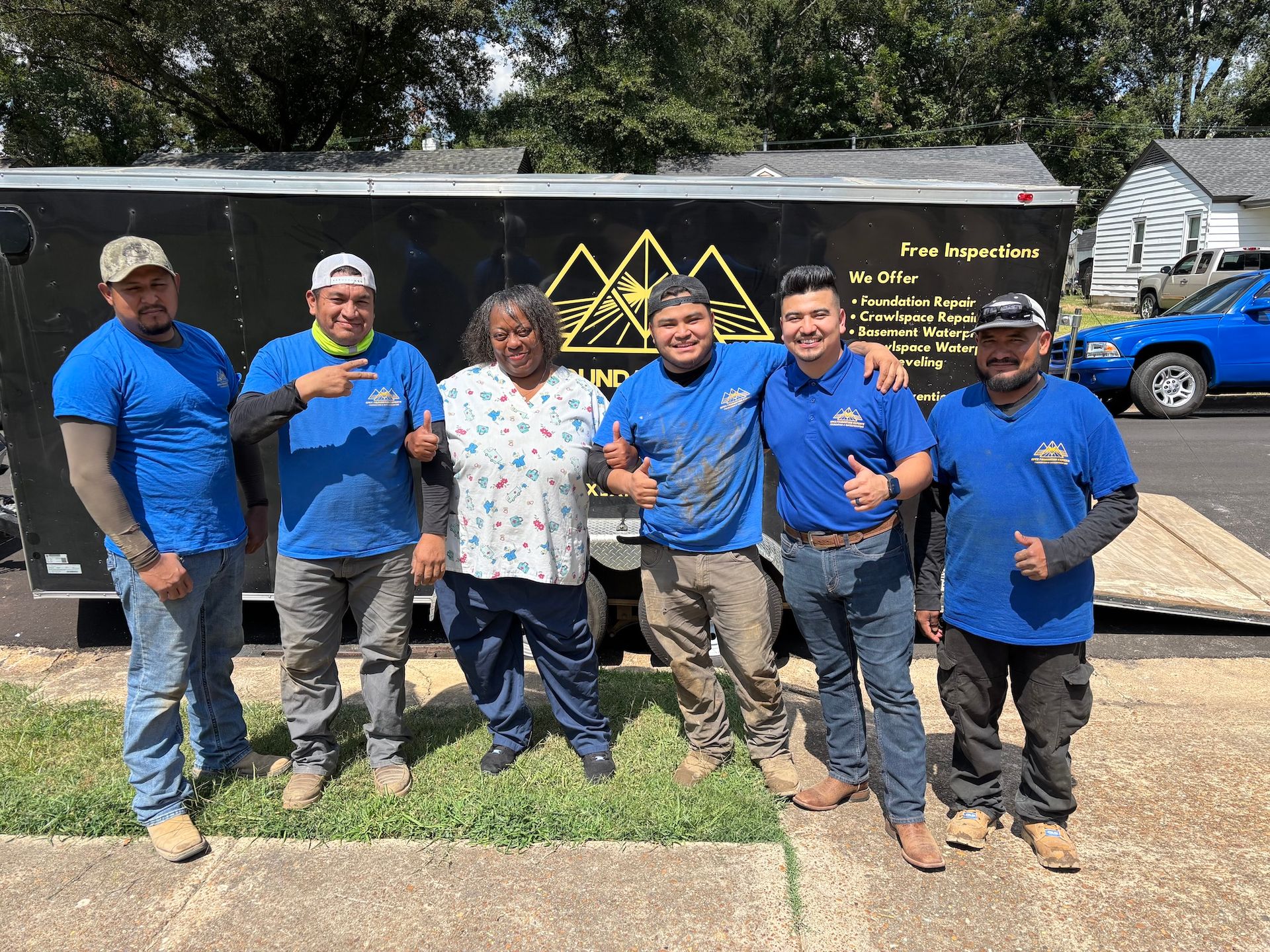 Six people wearing blue shirts stand in front of a dark construction trailer on a sunny, paved day.