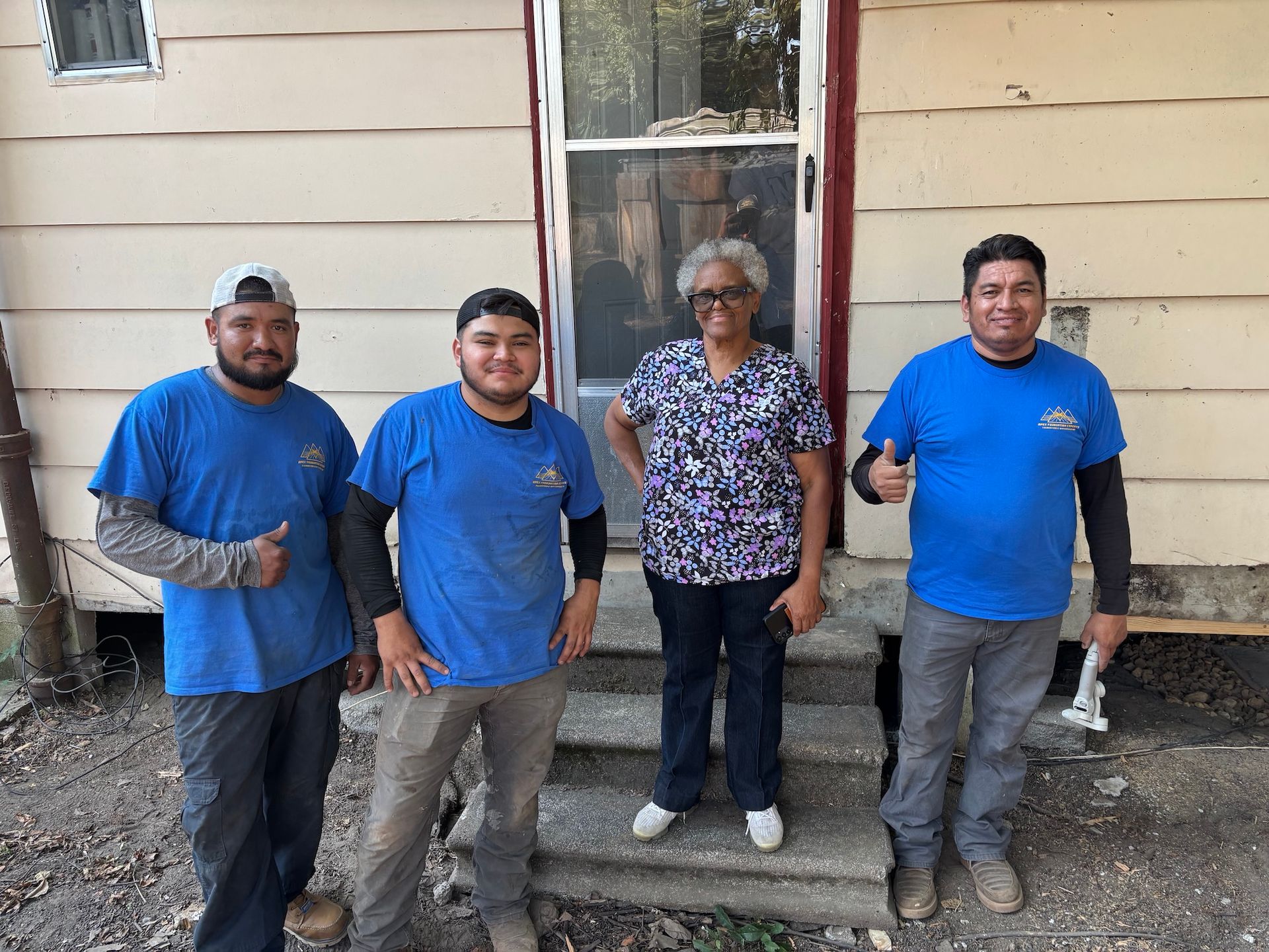 Four people posing in front of a house, three wearing matching blue t-shirts, with one person giving a thumbs up.