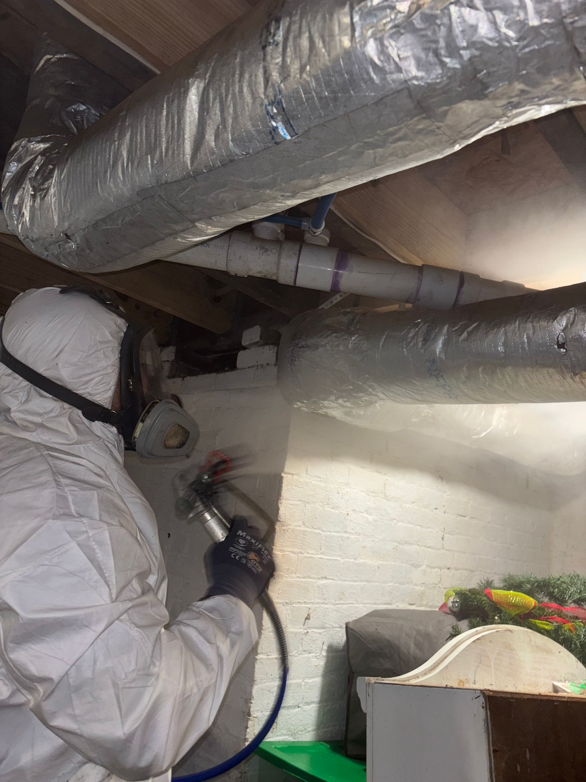 A person in protective gear sprays a white wall in a basement with exposed pipes overhead.