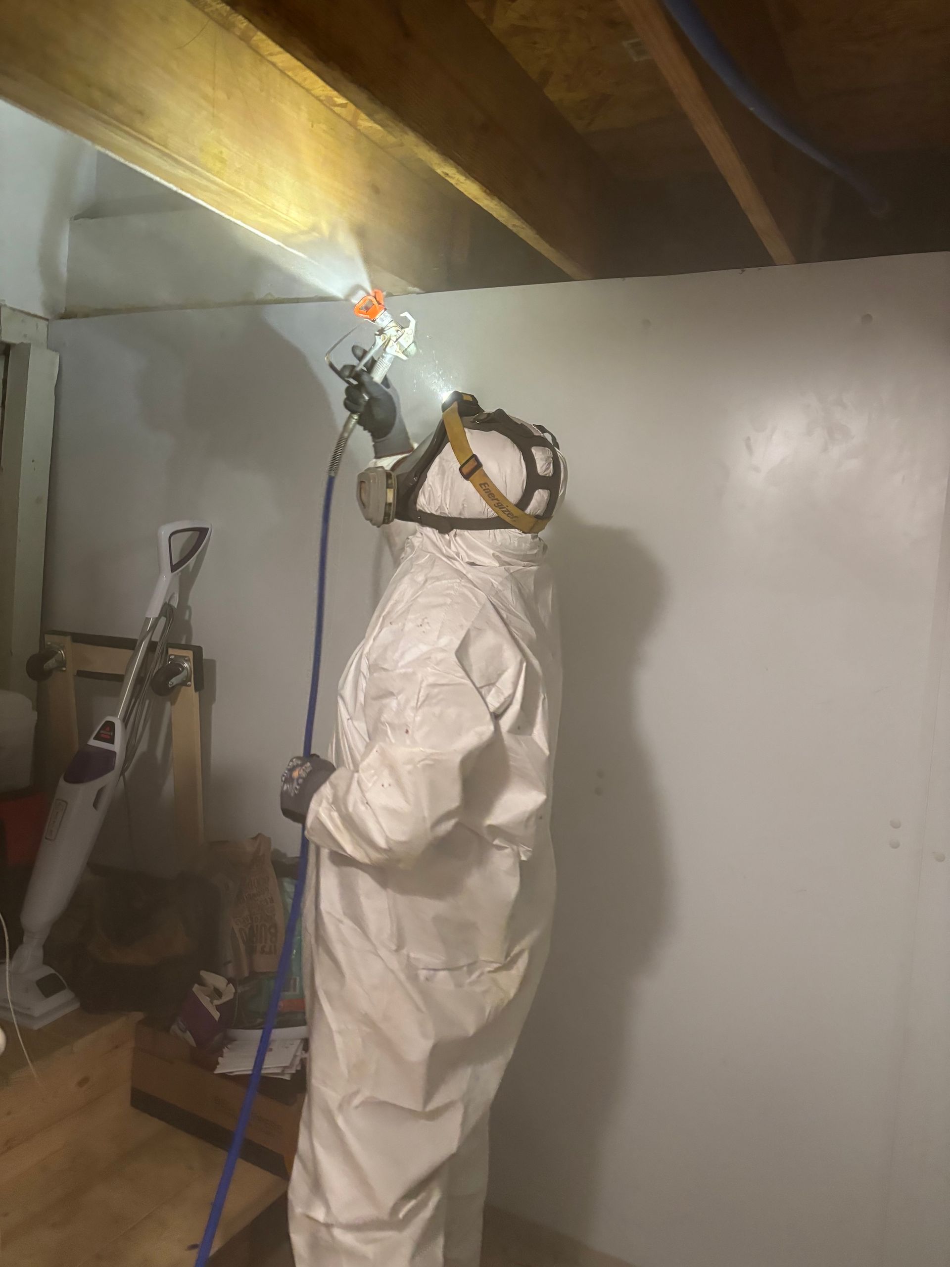 A person in a protective white suit and respirator uses a spray gun to paint a white wall in an unfinished room.