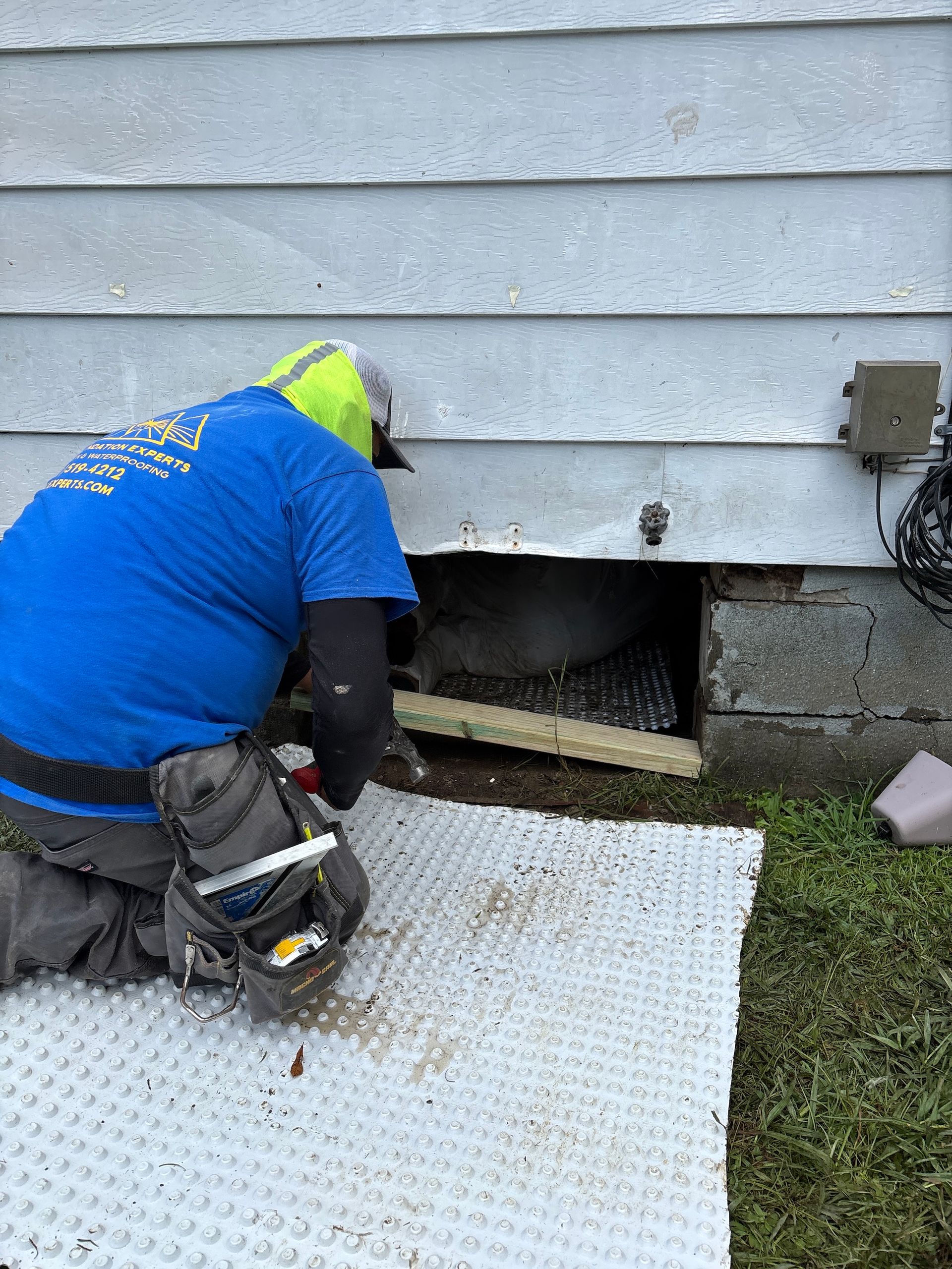 A technician in a blue shirt kneels on a mat, installing a barrier into an opening in a home's exterior foundation.