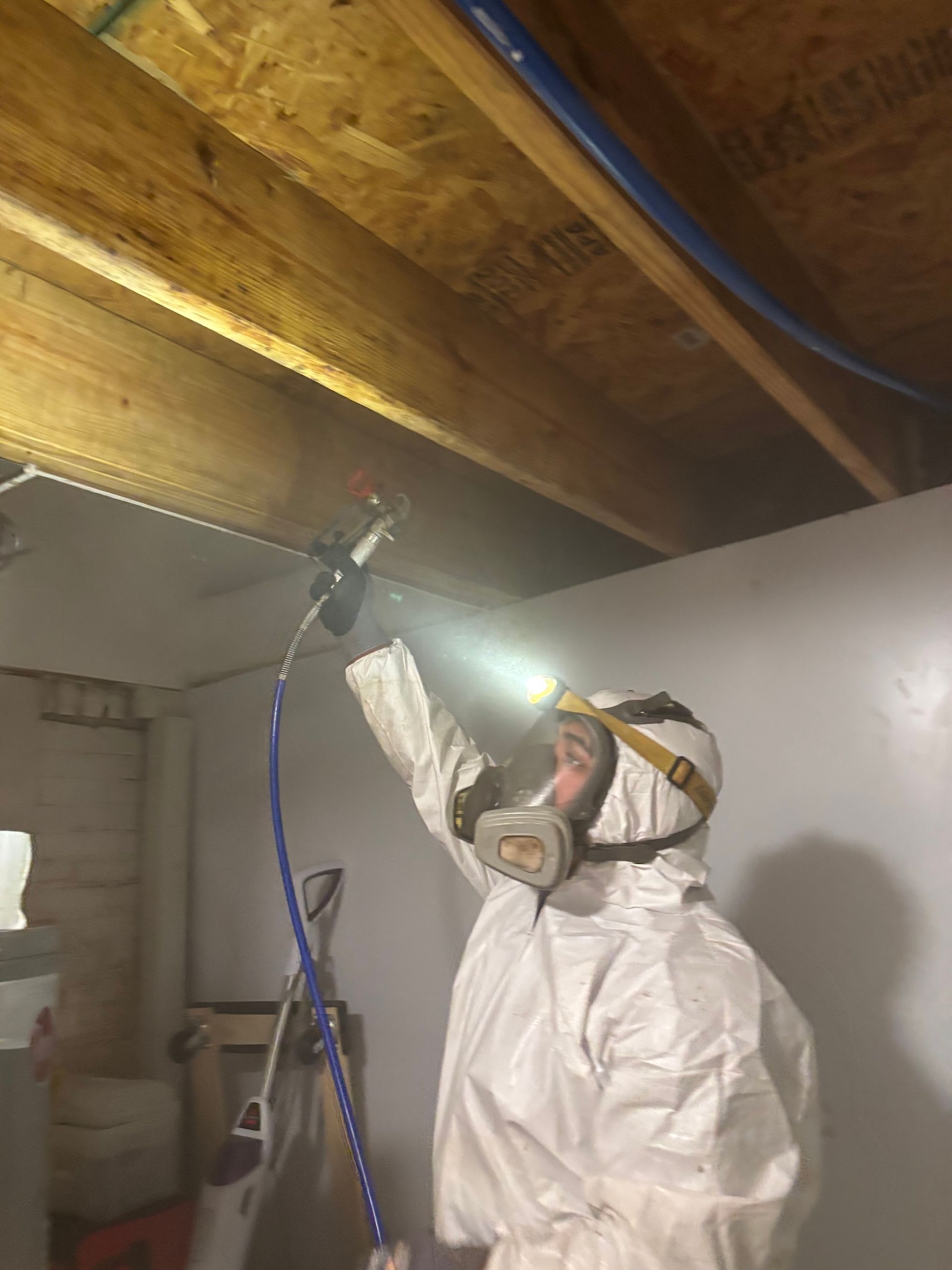 A worker in protective white coveralls and a respirator sprays a wooden beam in a basement using a handheld nozzle.