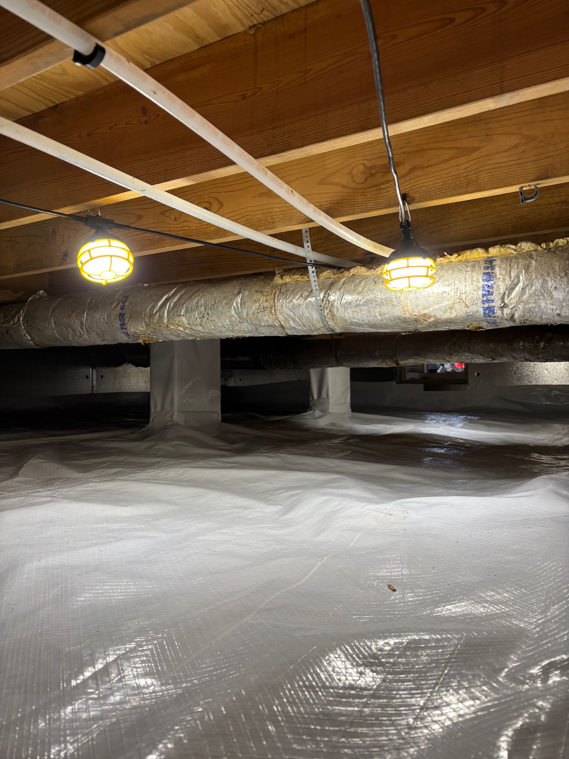 A crawl space with a plastic vapor barrier covering the dirt floor, support piers, ductwork, and hanging work lights.