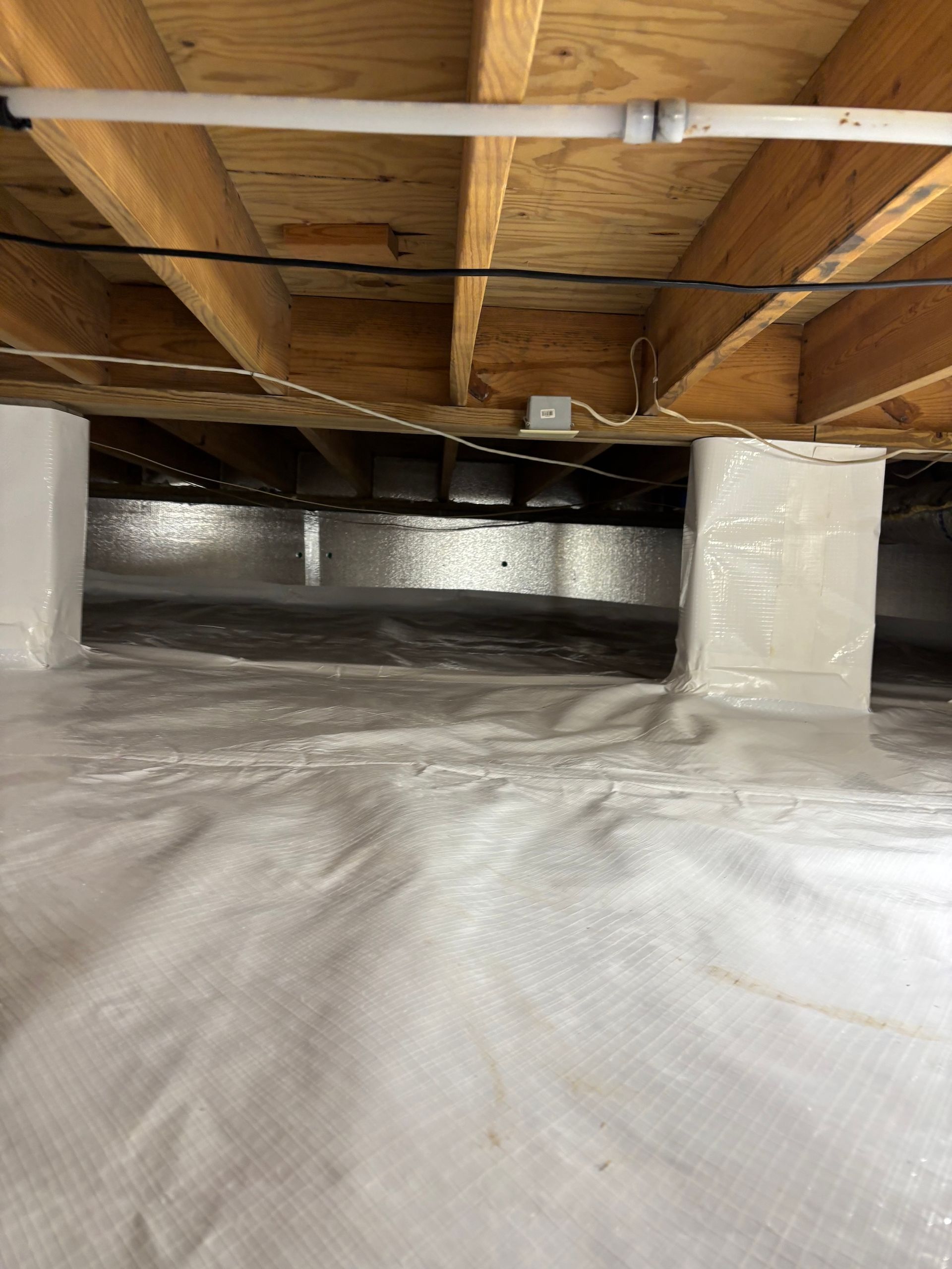 A crawl space with a clean white vapor barrier on the ground and white-wrapped structural piers supporting wood floor joists.