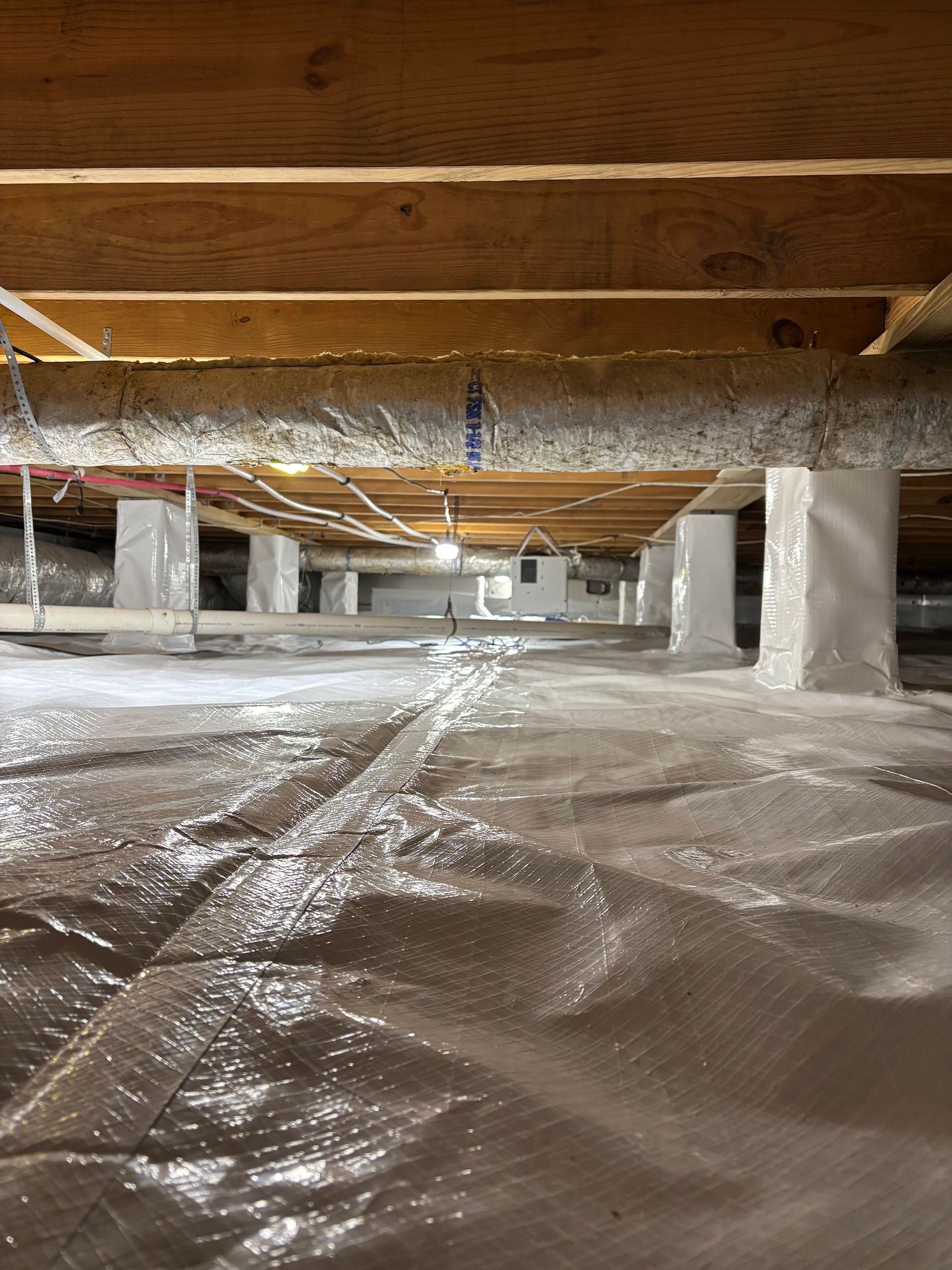 A crawl space featuring a clean white vapor barrier covering the ground, wooden joists, and white-wrapped support pillars.