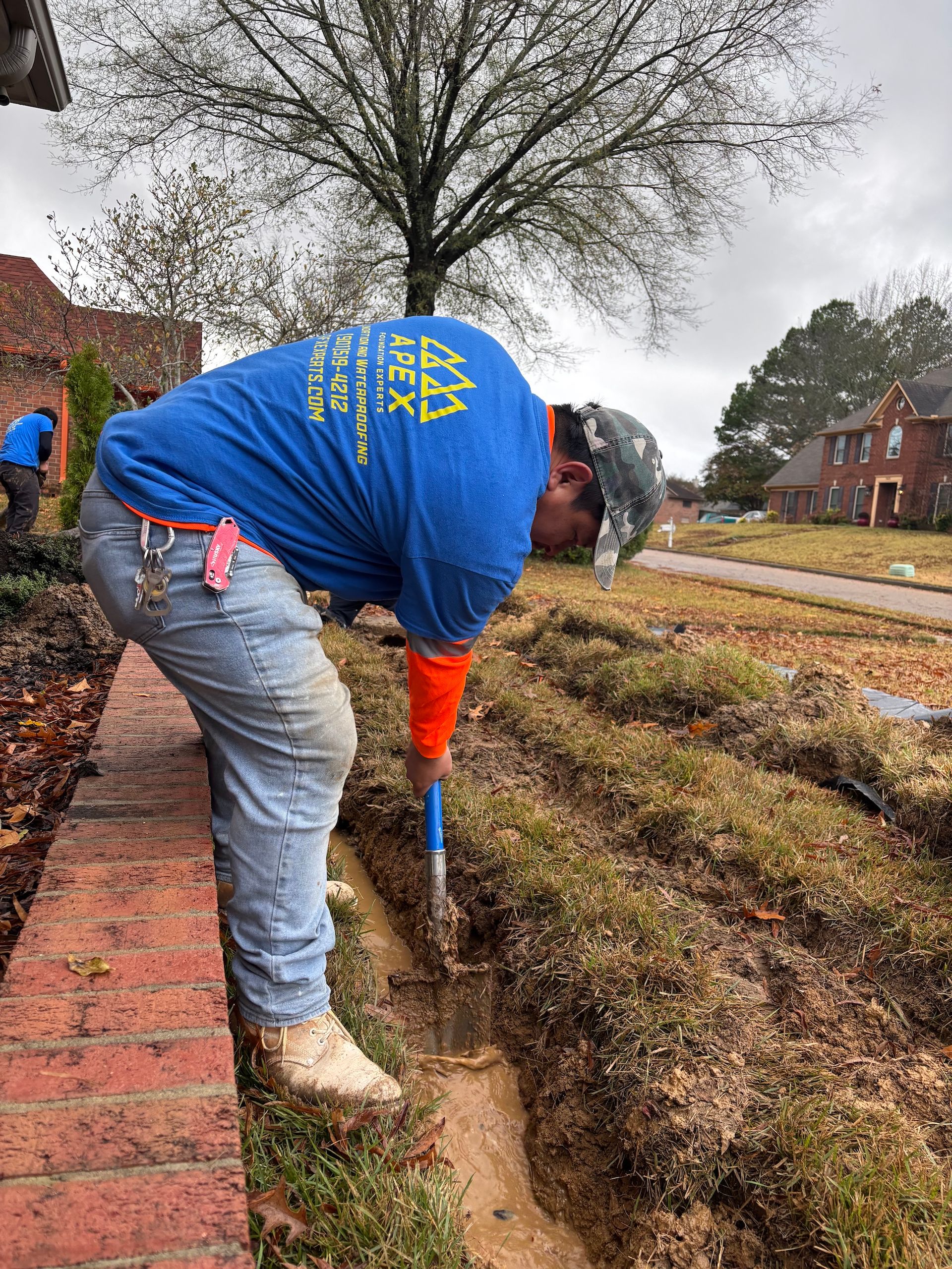 A worker in a blue t-shirt uses a shovel to dig a trench along a brick wall in a residential yard.