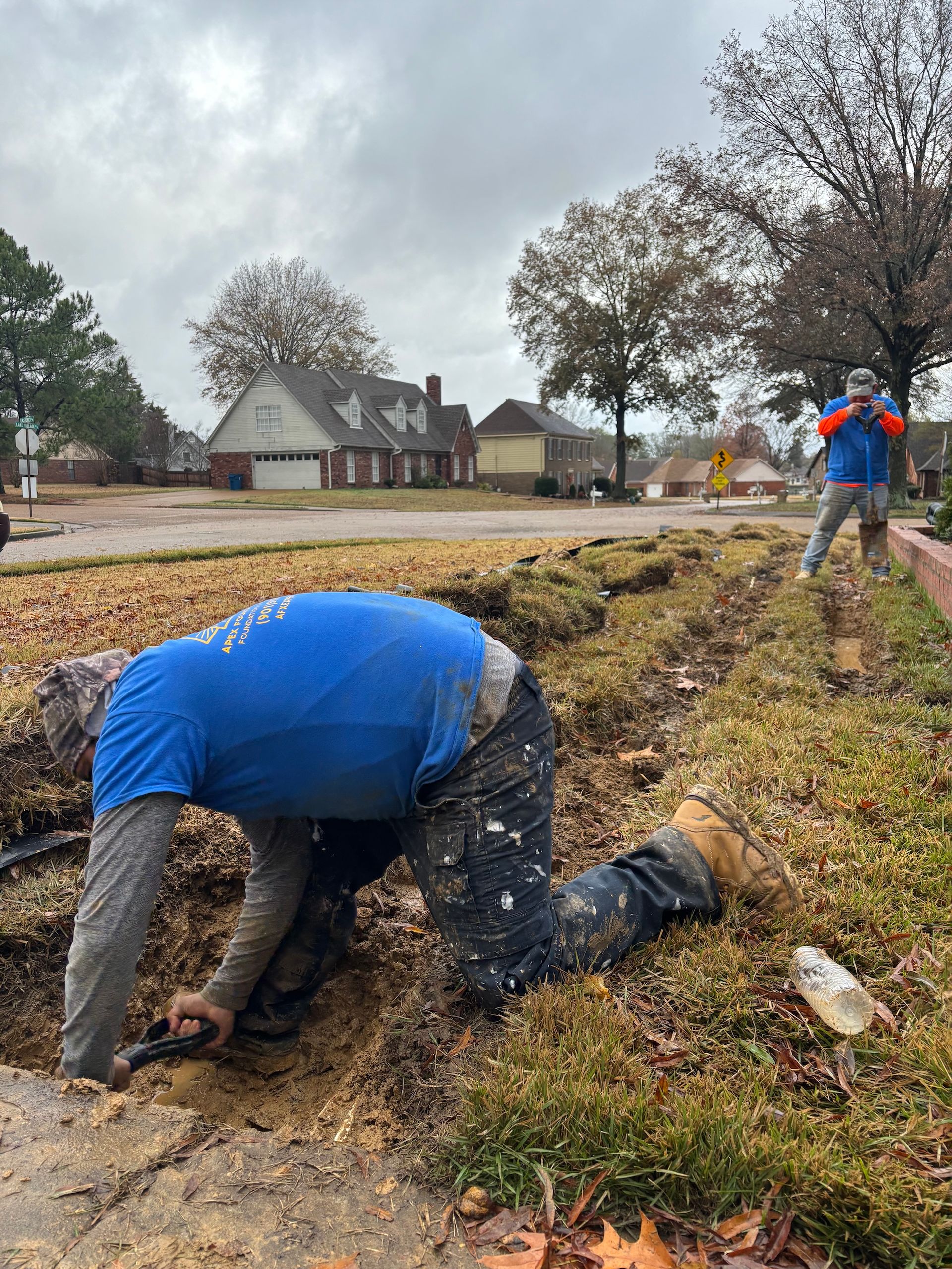 Two people in blue shirts work on a landscaped yard, with one kneeling to dig in the soil near a sidewalk.