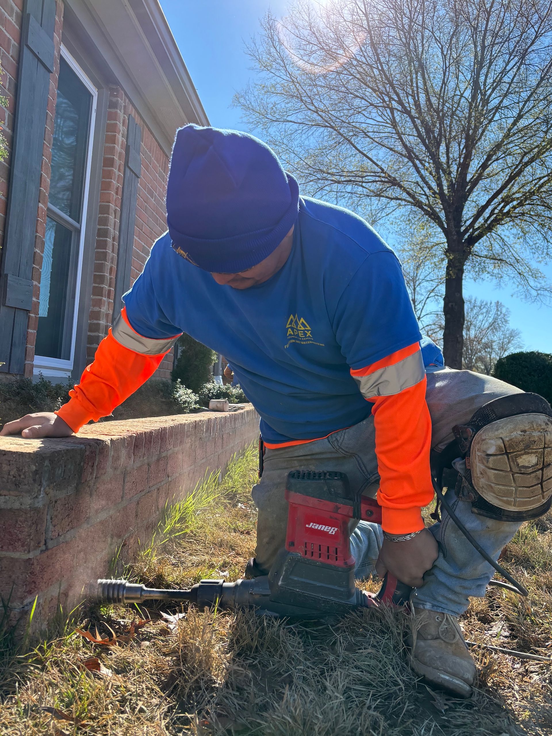 A person wearing a beanie and high-visibility sleeves uses a power tool to break apart a brick retaining wall.