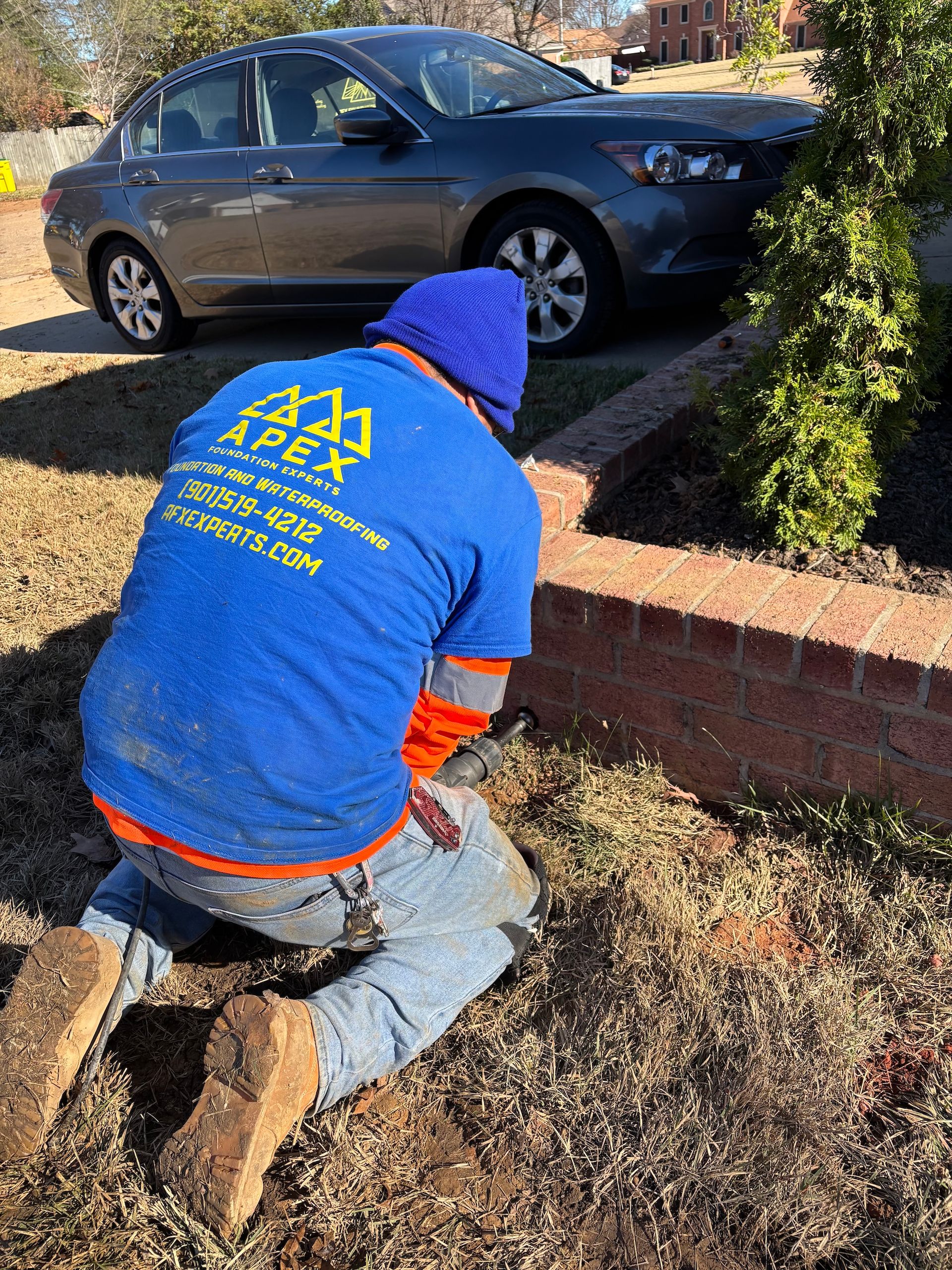 A worker in a blue Apex uniform kneels on the grass, performing maintenance near a brick planter with a car in the back.