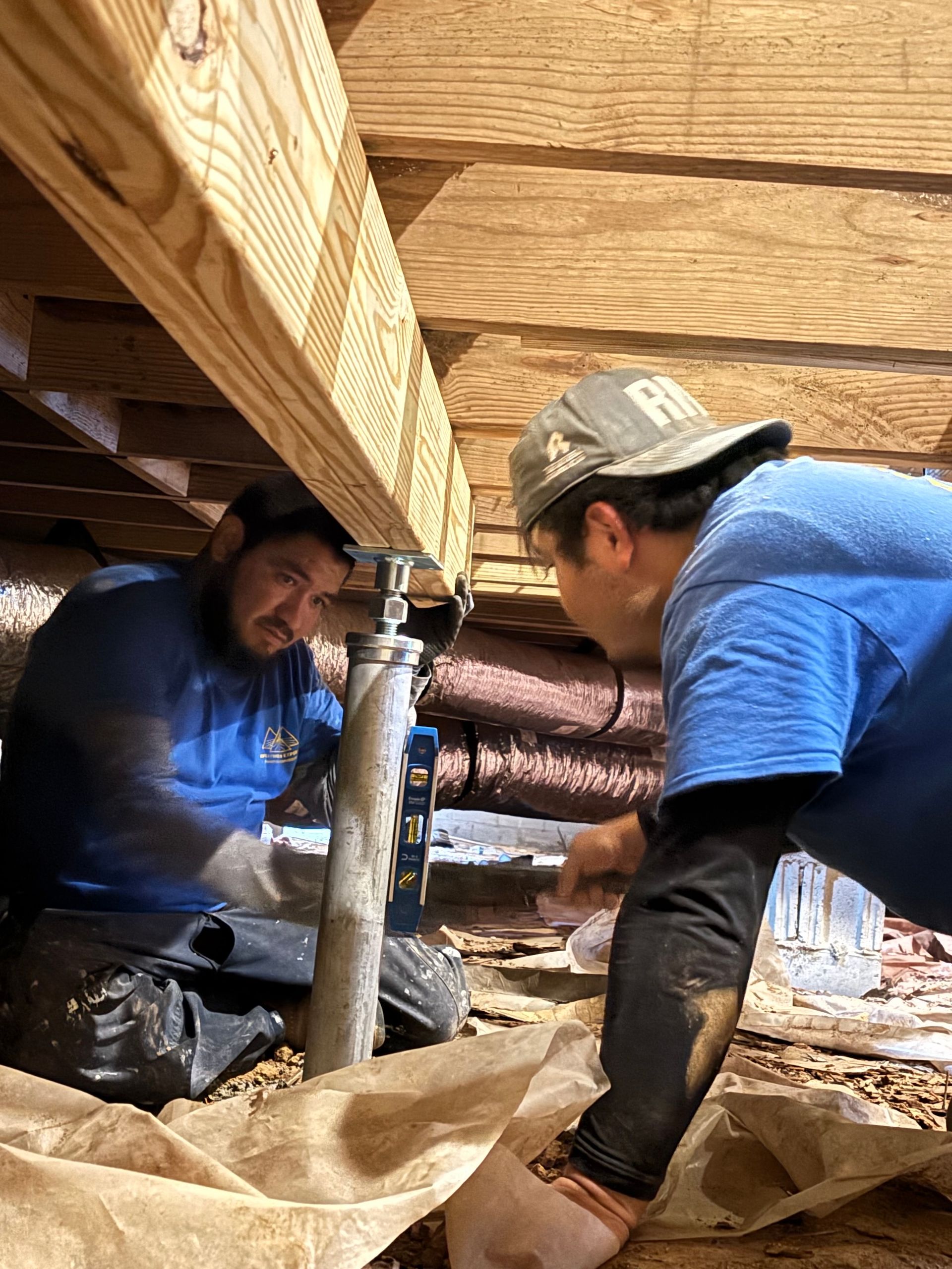 Two workers in a crawl space install a metal support jack under a wooden floor joist.