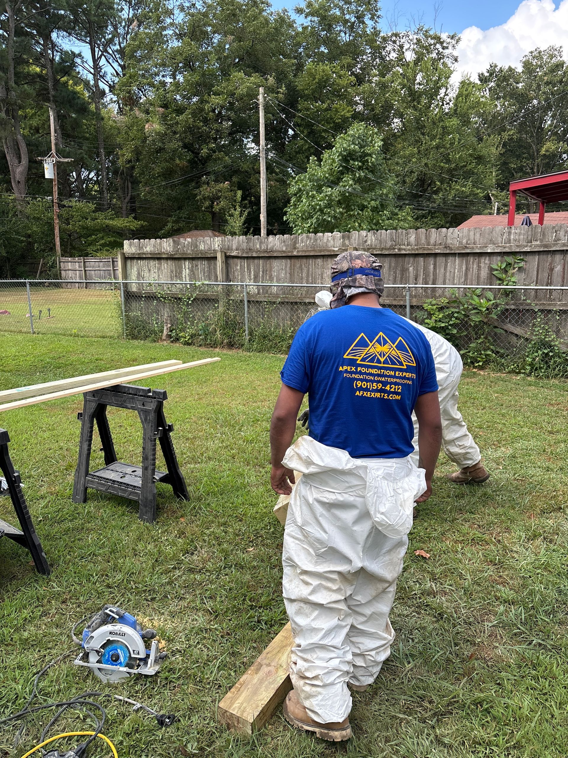 Two people in white protective coveralls work on a wooden board in a backyard, using a circular saw on a sawhorse.