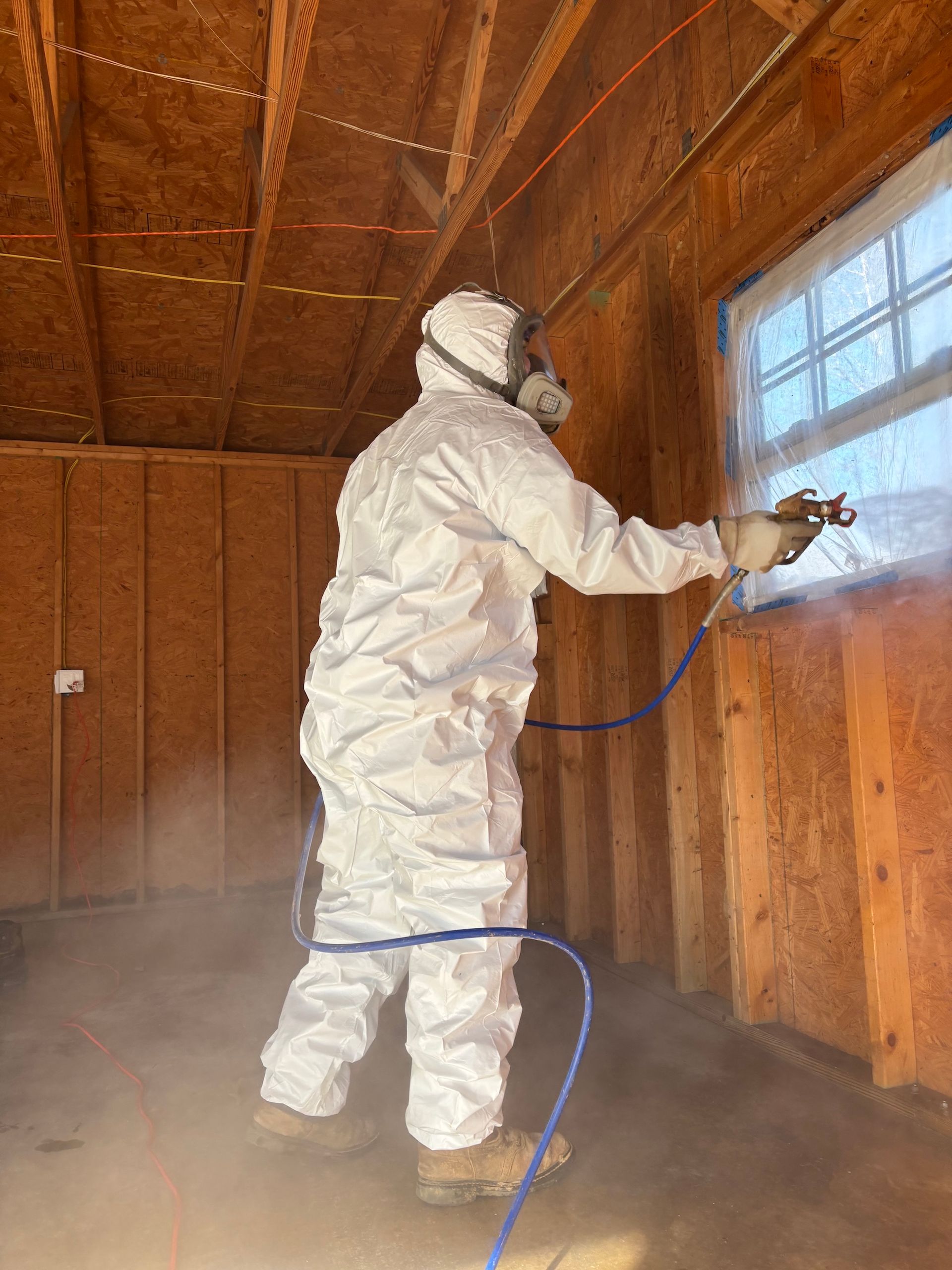 A person in a full white protective suit and respirator sprays a substance onto a wall in an unfinished wooden building.