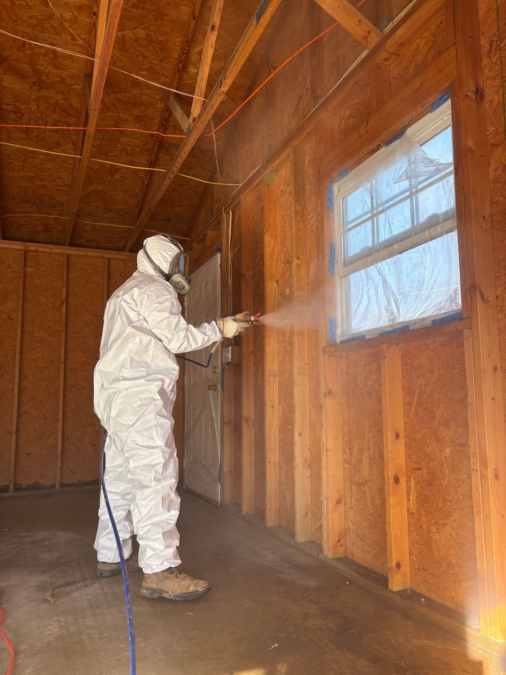 A person in protective white gear uses a spray tool to apply coating to the interior wood walls of a structure.