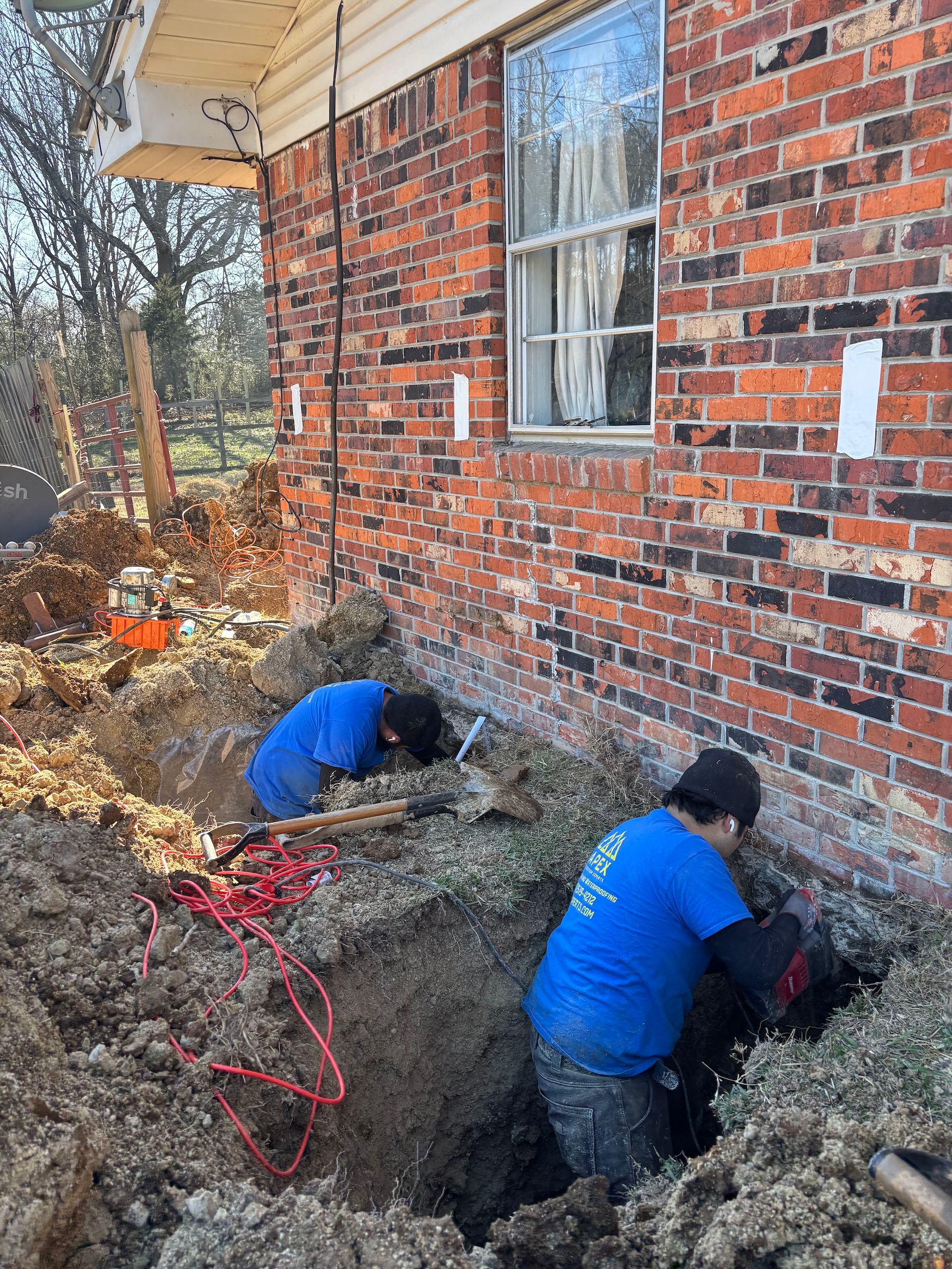 Two workers in blue shirts dig trenches along a brick residential foundation to install structural supports.