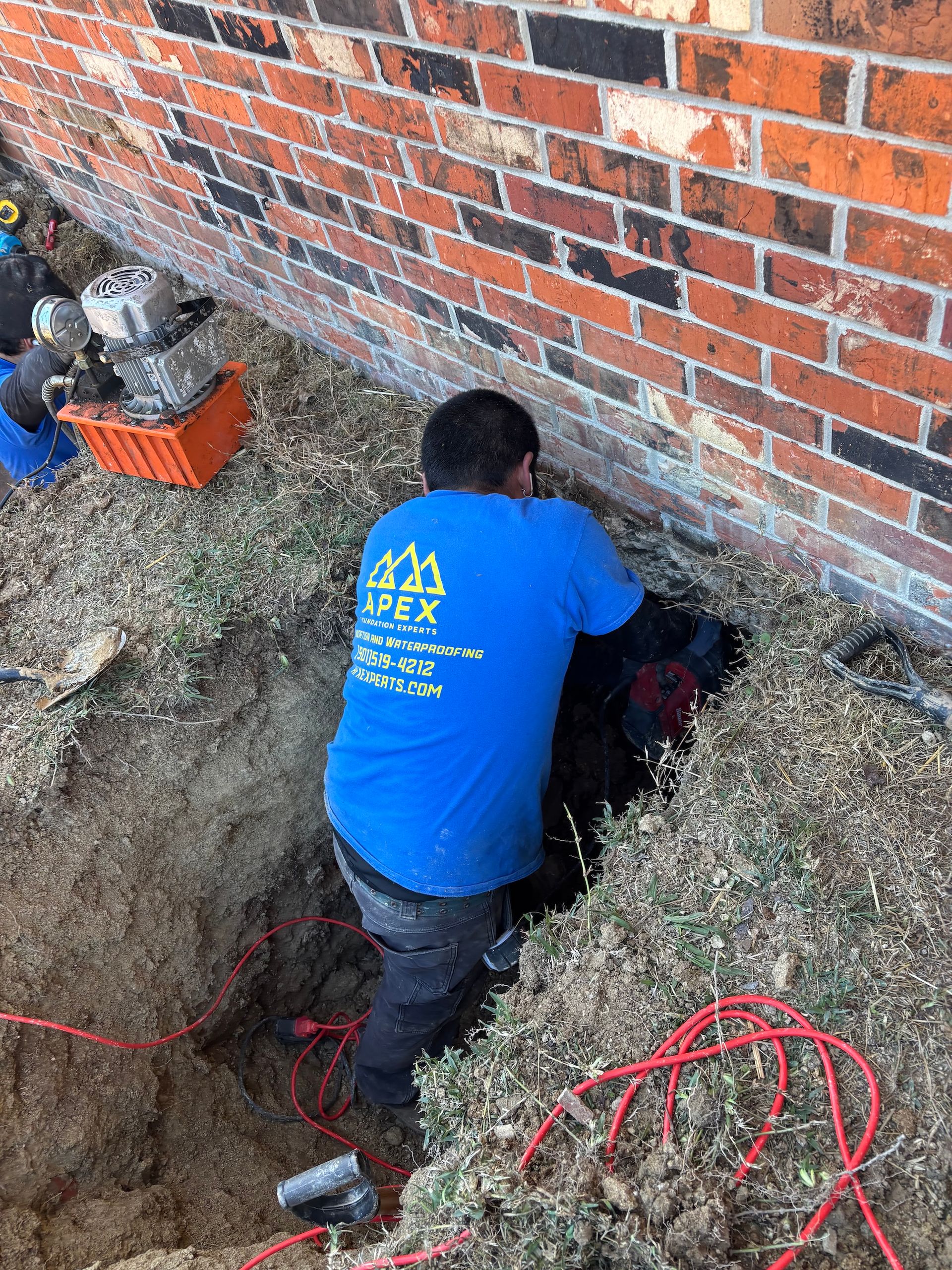 A technician in a blue Apex shirt works in a hole dug along the exterior brick wall of a building.
