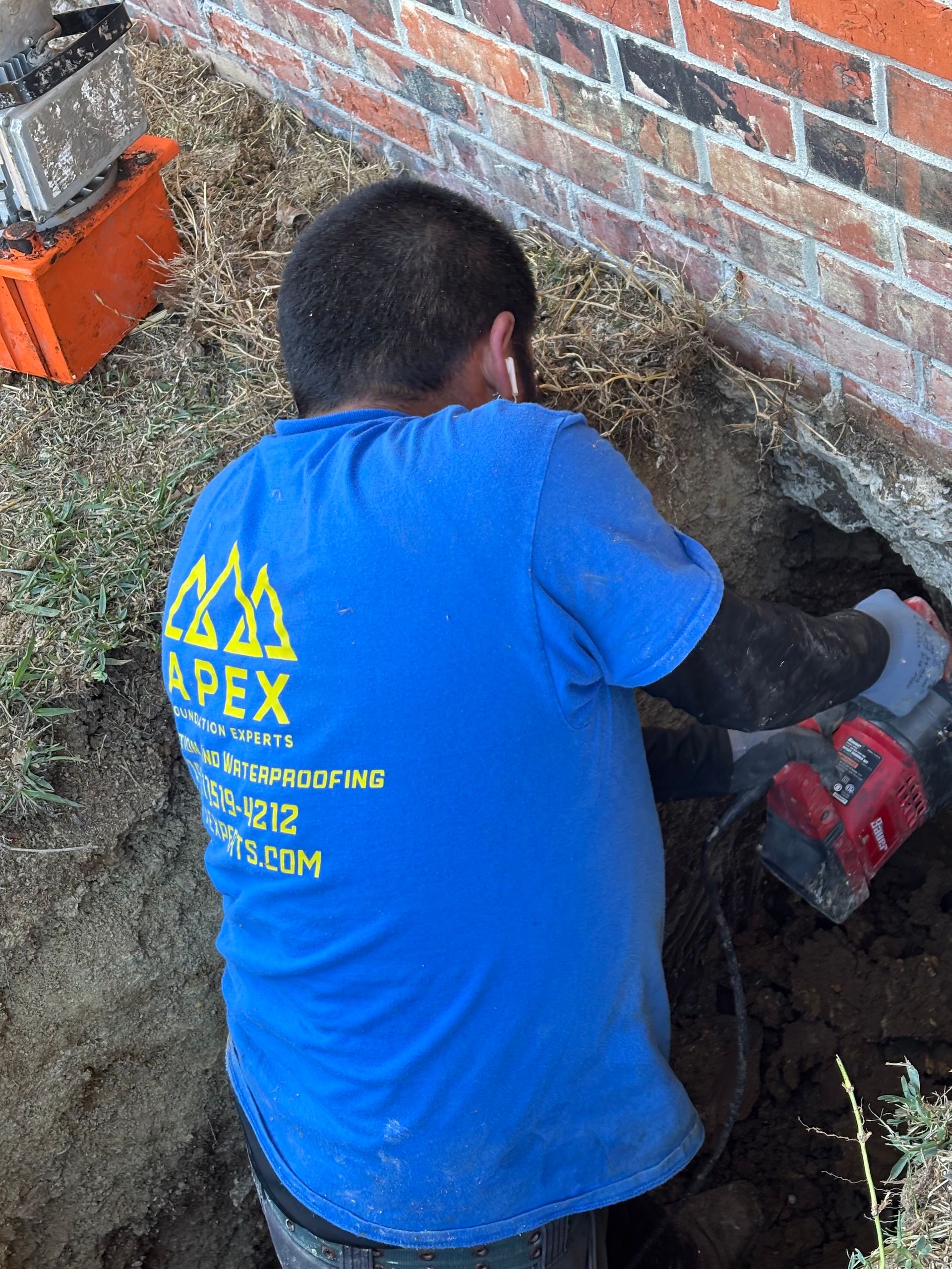 A worker in a blue Apex shirt kneels by a brick wall, using a red power tool to dig into the ground.